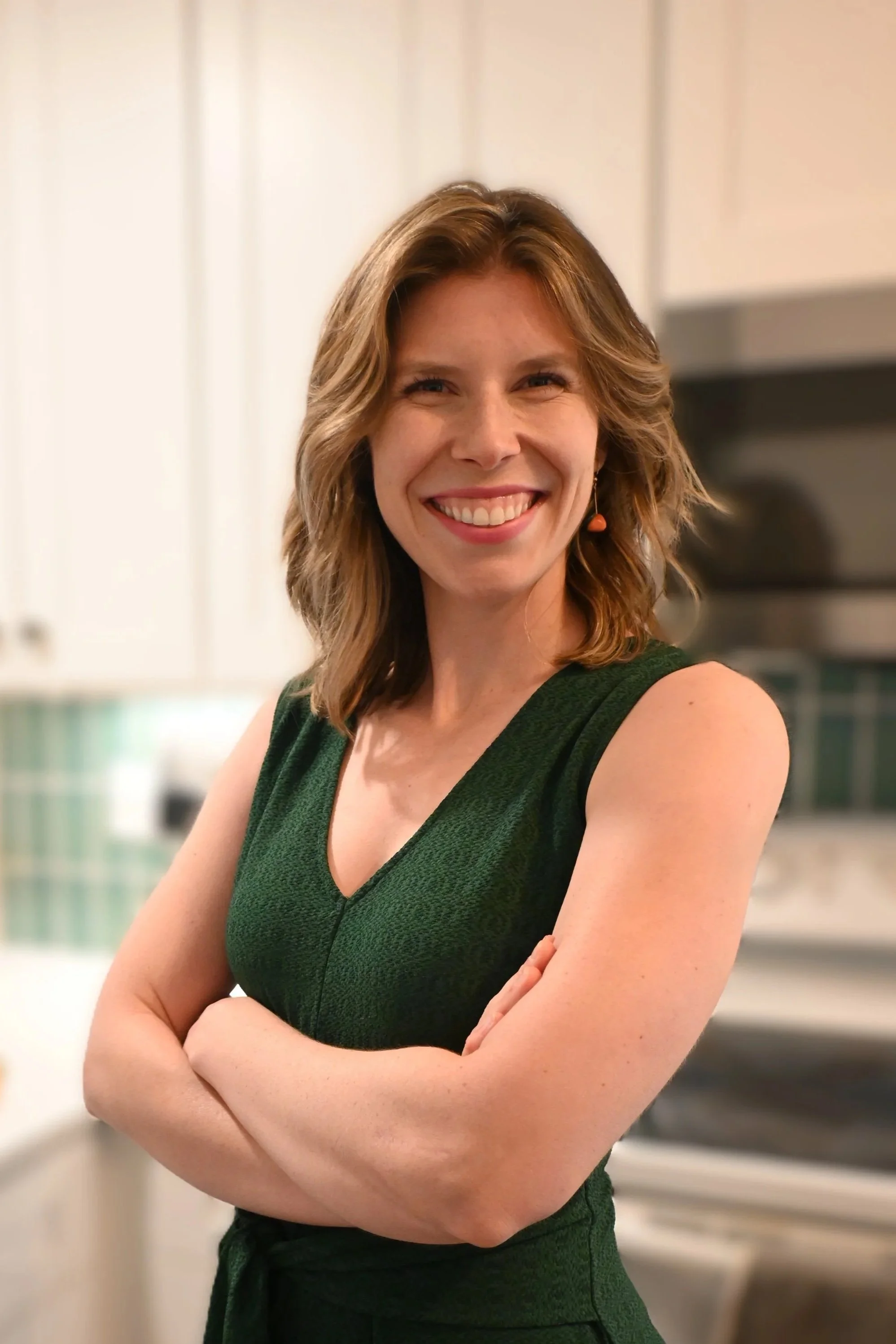 A woman with shoulder-length wavy hair smiling, wearing a sleeveless green dress, standing with arms crossed in a kitchen.