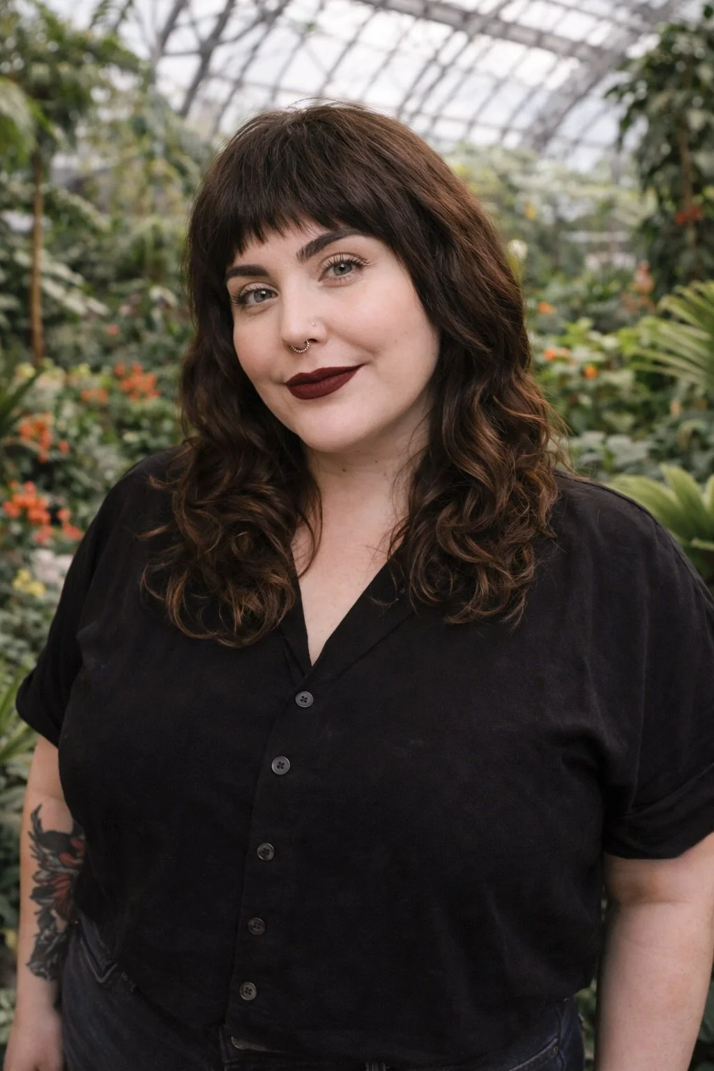 A woman with dark brown wavy hair, dark lipstick, a septum piercing, wearing a black shirt, standing in a greenhouse with green plants and orange flowers in the background.