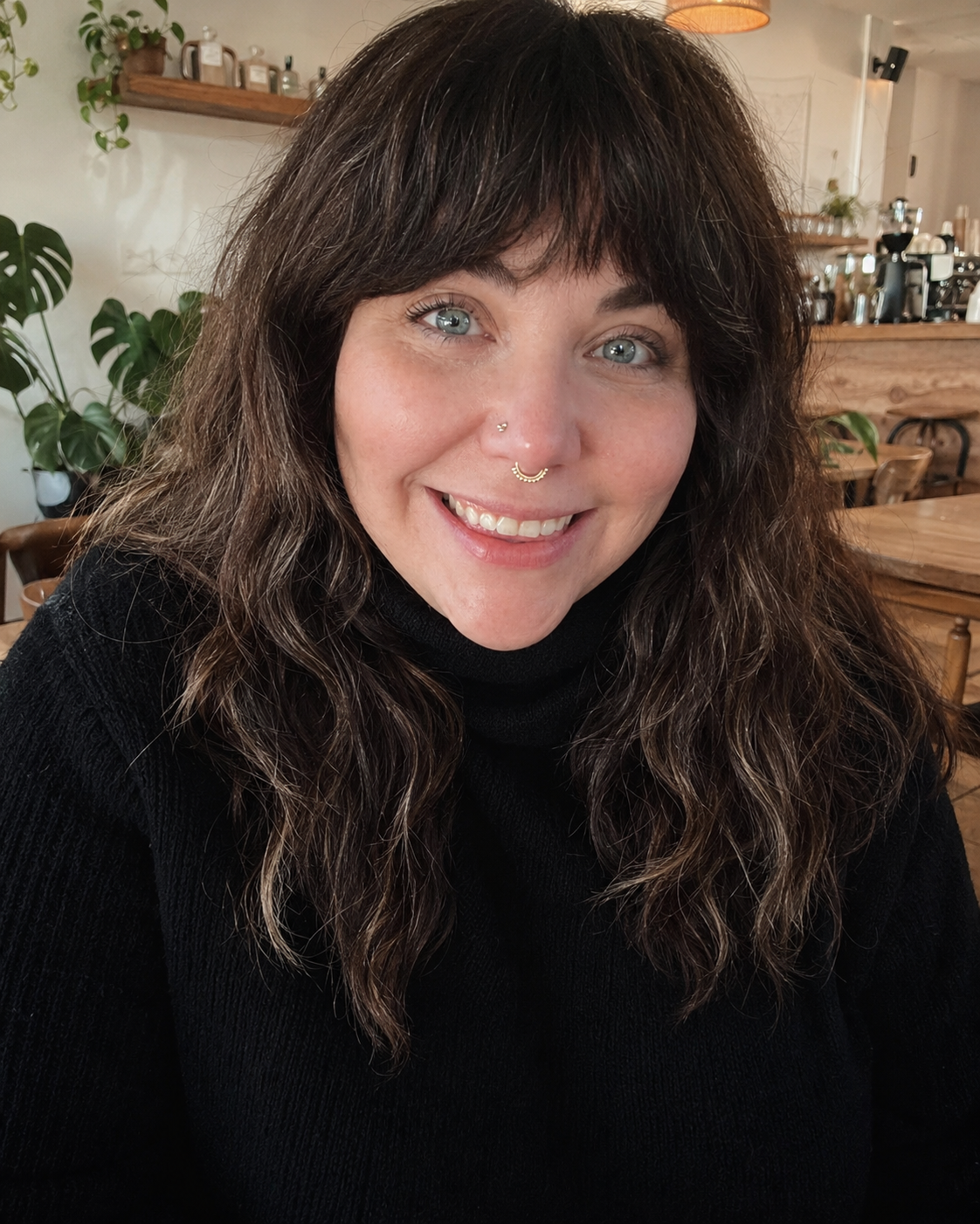 A smiling woman with wavy brown hair and blue eyes, wearing a black turtleneck, sitting in a cozy cafe with wooden furniture and green plants in the background.