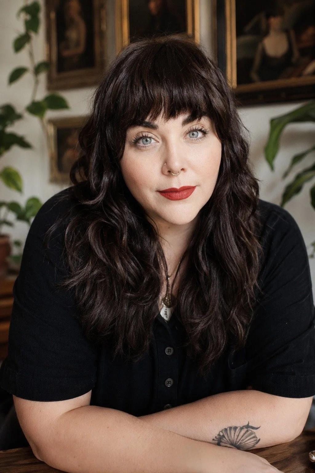 A woman with dark wavy hair, blue eyes, and red lipstick looking at the camera while sitting at a table with her arms crossed. She is wearing a black shirt and has a tattoo of a flower on her forearm. The background shows framed paintings and green plants.