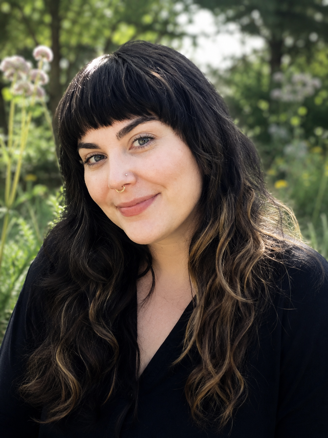 A woman with long, wavy brown hair with blonde highlights, light skin, and a septum piercing, smiling outdoors with green trees and pink flowers in the background.