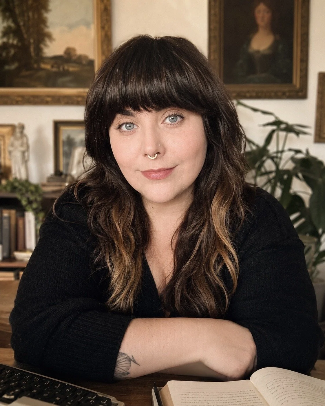 A woman with dark brown, wavy hair, light skin, and blue eyes sitting at a table with an open book and a keyboard in front of her. She has a septum piercing and is wearing a black top. The background features framed art and a bookshelf.