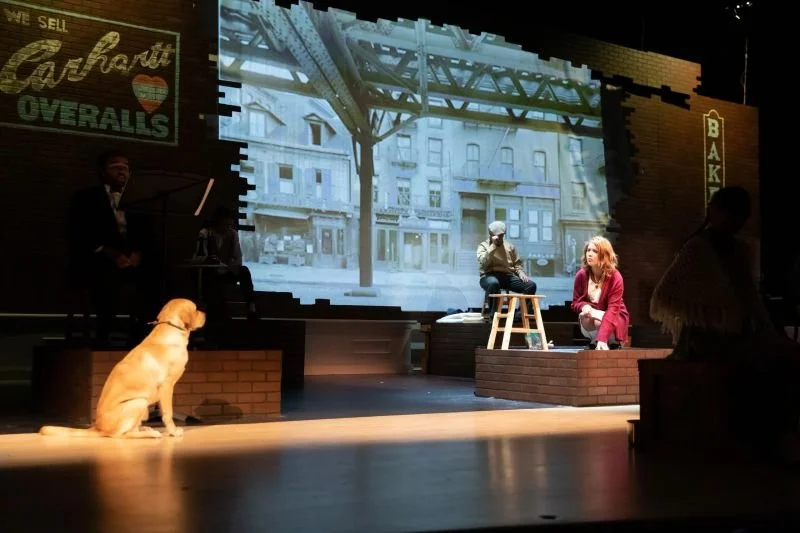A stage performance with two actors sitting on a brick platform and stool, with a large screen behind them displaying a city street scene. There are stage lights shining on the scene, and a dog is sitting on the floor in the foreground.