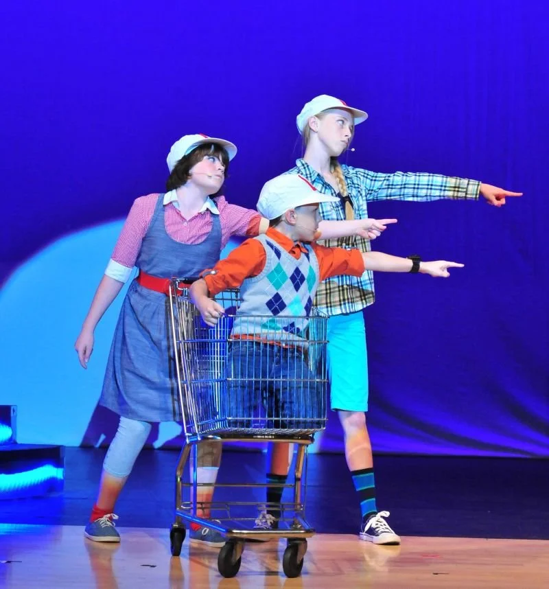 Three children on stage performing a play, with one in a shopping cart, all wearing casual clothes and white caps, pointing in different directions against a blue background.