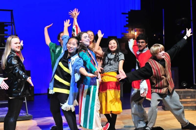 Group of children on stage performing in colorful costumes with a blue backdrop.