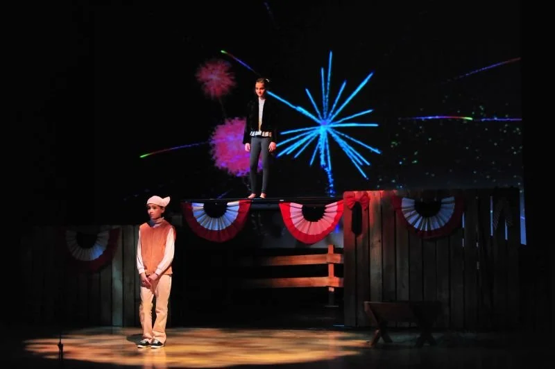 Two children performing on a stage with patriotic red, white, and blue bunting, one standing on the stage and the other standing on a raised platform, with a background projection of fireworks.