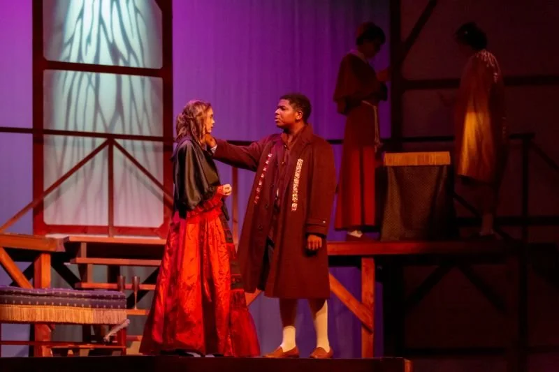 Two actors on stage, a woman in a red skirt and dark jacket and a man in a long brown coat, engaging in a scene. Two other women are in the background, one standing on a wooden platform and the other near a table, both in period costumes. The scene h
