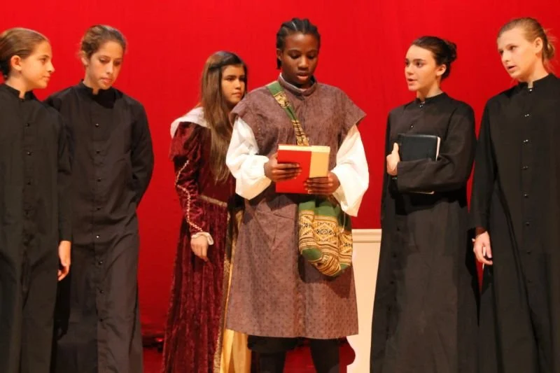 Group of five young girls in costumes standing on stage with a red curtain background, participating in a play or performance.