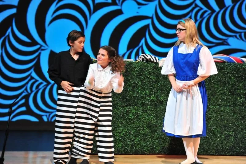 Three young girls performing on stage with a black and blue swirling patterned backdrop. Two girls are dressed in black and white striped pants, and one girl is wearing a colonial-style dress. They are engaged in a theatrical scene.