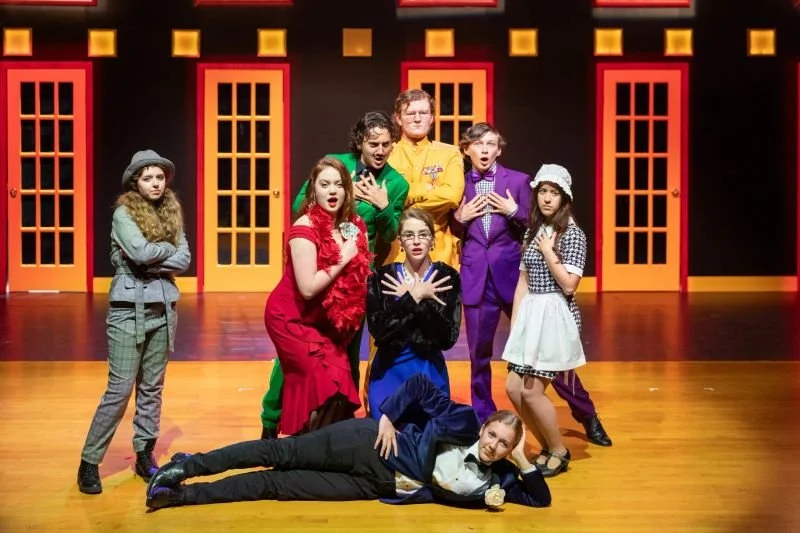 Group of children in colorful costumes posing on a stage with a set resembling a neighborhood or cityscape in the background.