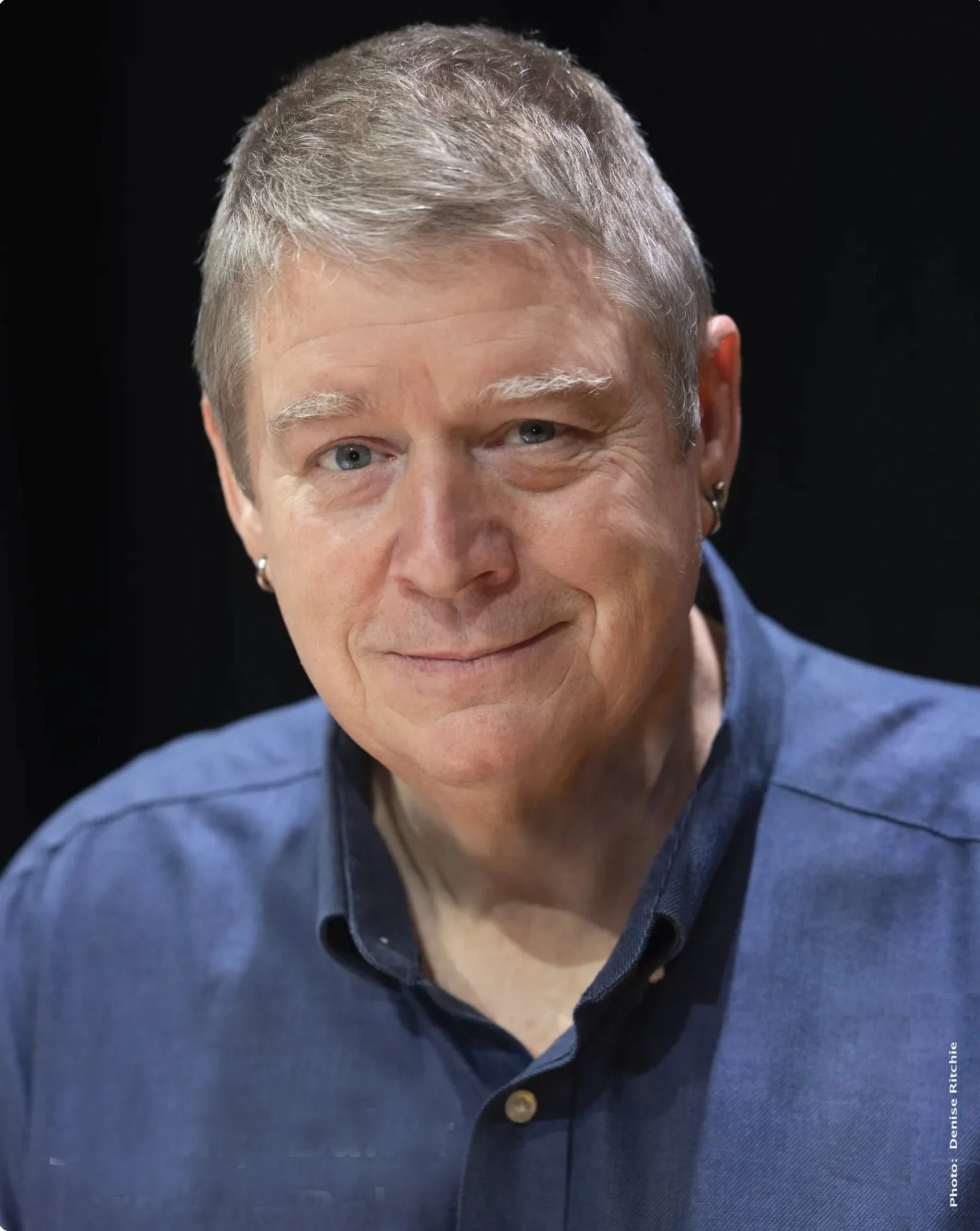 Close-up of a man with short gray hair and blue eyes, wearing a blue collared shirt, smiling against a black background.