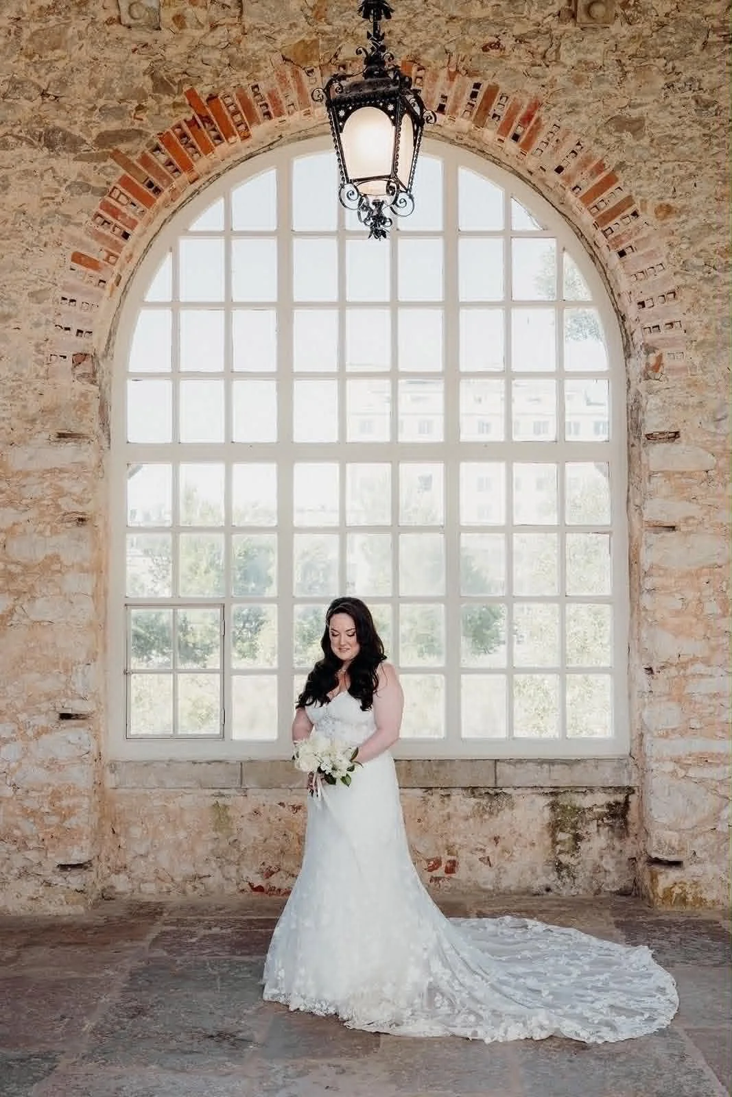 No interior de uma sala com parede de pedra, uma mulher em vestido de noiva segura um buquê de flores brancas na frente de uma grande janela de vidros, com um lustre de ferro antigo pendurado no teto.