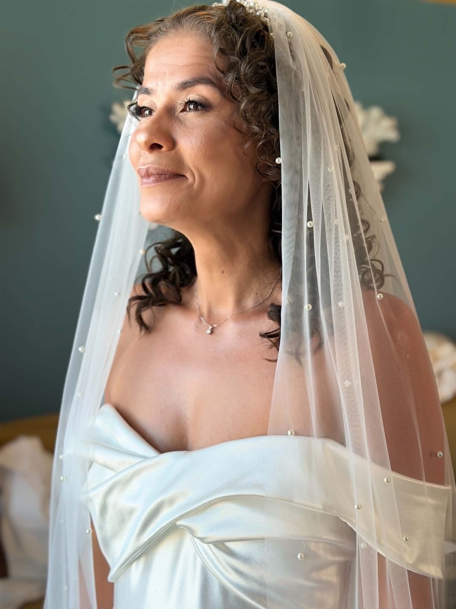 Mulher com cabelo cacheado usando véu de noiva de tule com pérolas, sorrindo suavemente.