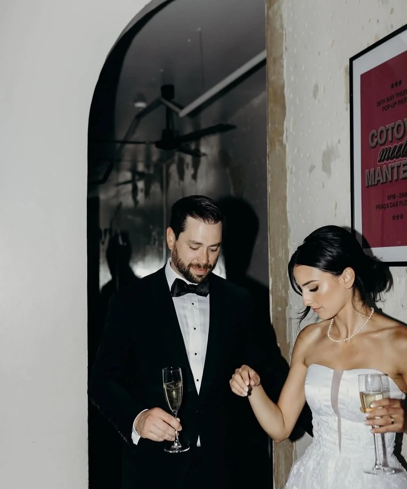 Homem vestido de terno preto e gravata borboleta e mulher de vestido branco elegante com colar de pérolas em uma festa ou evento social, ambas segurando taças de espumante.