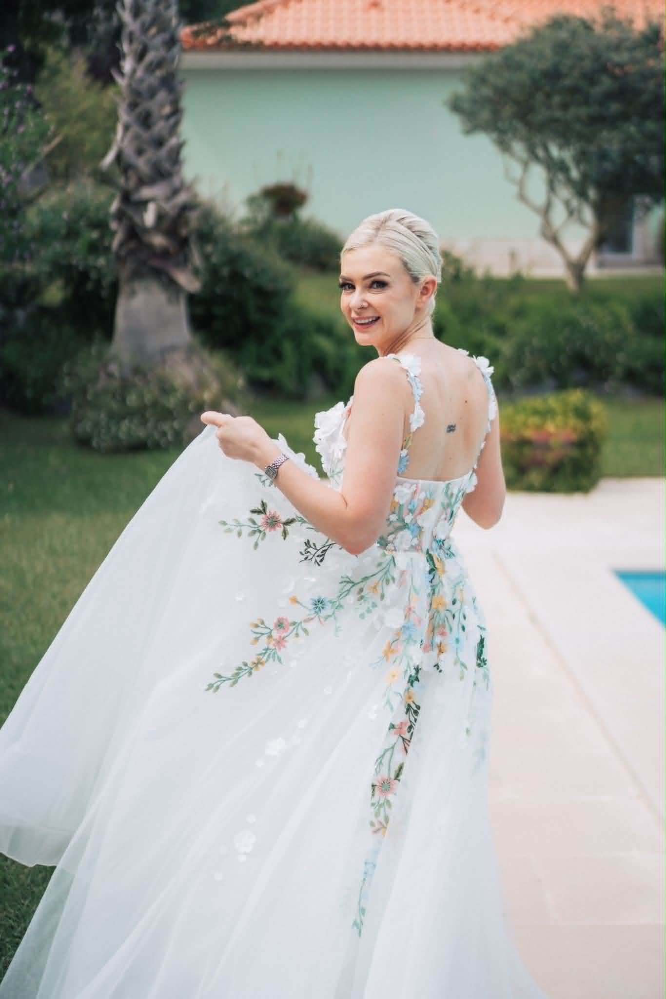 Mulher de cabelo platinado e vestido floral branco, sorrindo e voltando-se para trás na área ao ar livre com árvores e casa ao fundo.