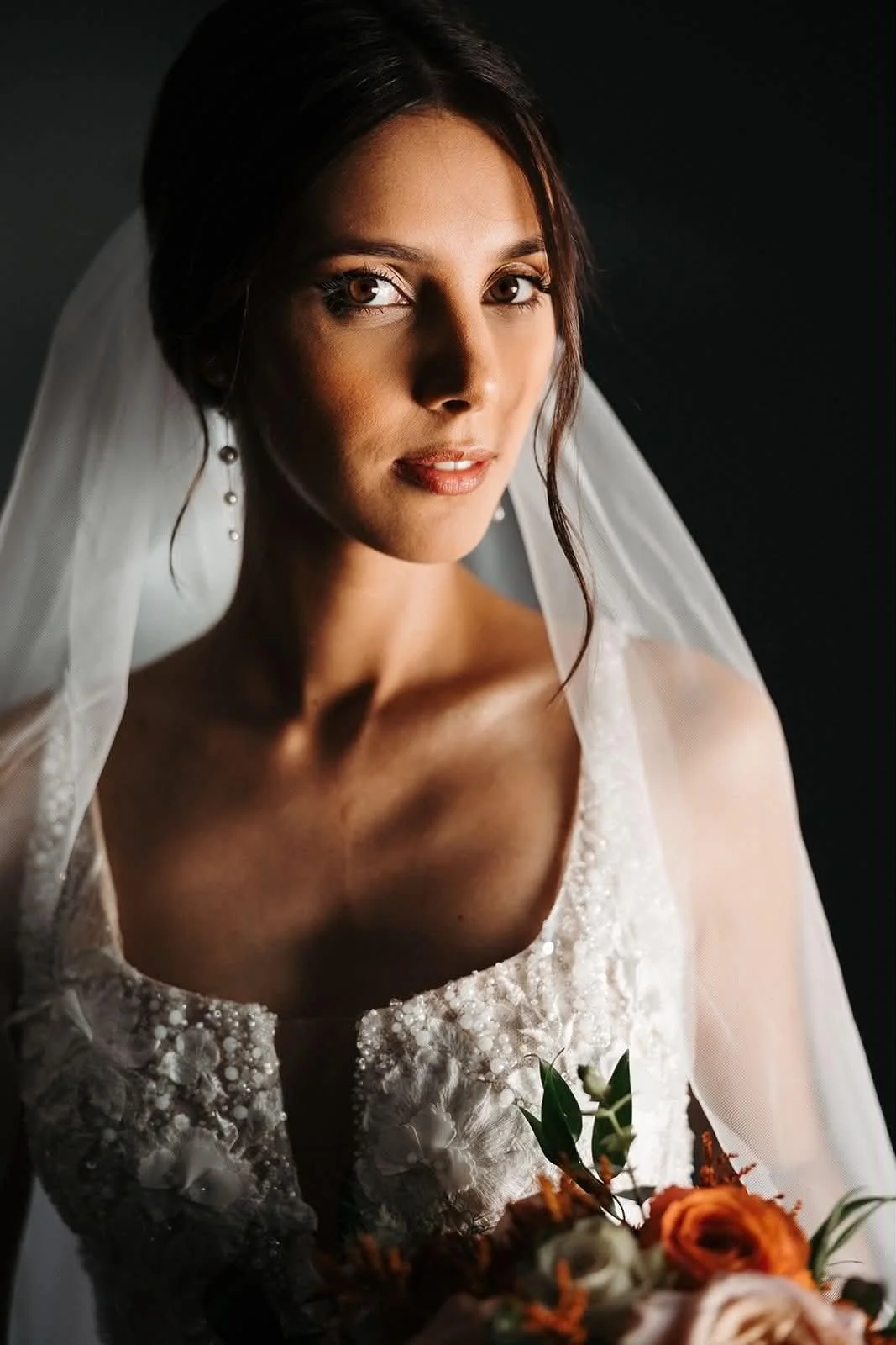 Mulher vestida de casamento com vestido branco, véu, brincos e bouquet de flores, olhando para a câmera com fundo escuro.
