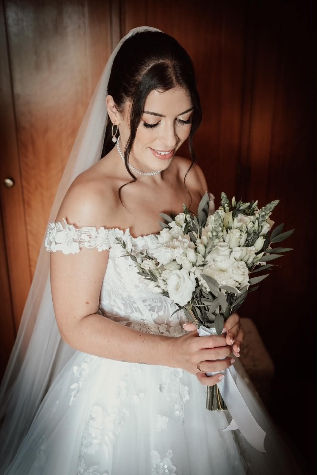Noiva sorridente com vestido branco, segurando um buquê de flores brancas e verdes, com cabelo preso e véu, em ambiente com paredes de madeira.