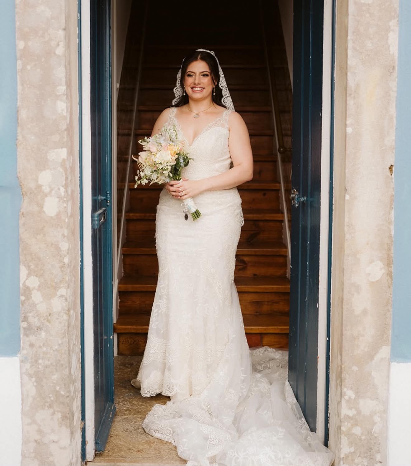 Noiva sorridente de vestido de casamento, segurando buquê de flores, posando na entrada de uma casa com escada de madeira ao fundo.