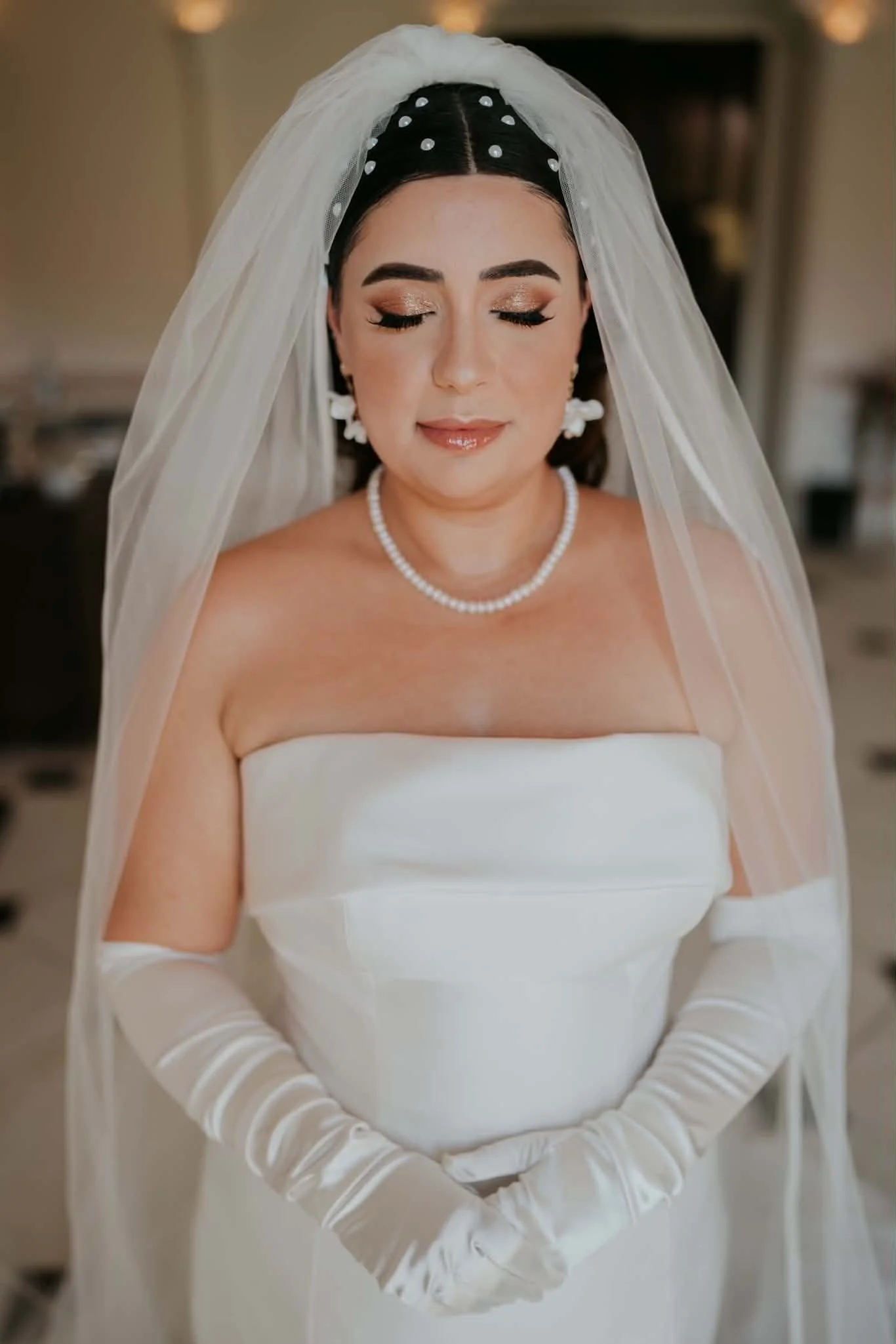 Mulher usando vestido de noiva branco, com véu, maquiagem elegante, brincos grandes, colar de pérolas e luvas brancas, com os olhos fechados.