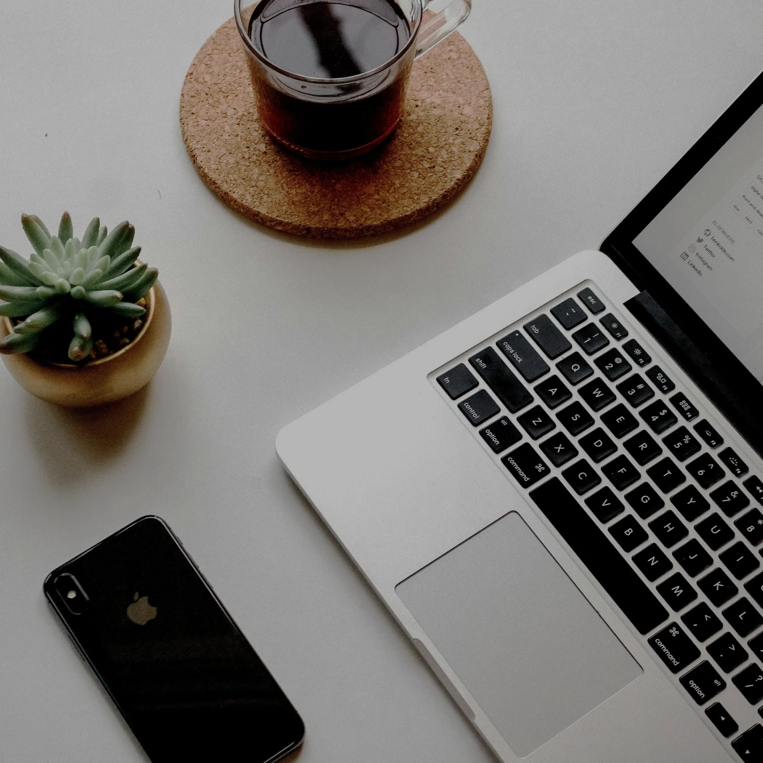 Top view of a workspace with a laptop, a smartphone, a glass cup of coffee on a cork coaster, a small potted succulent, and part of a digital device on a white surface.