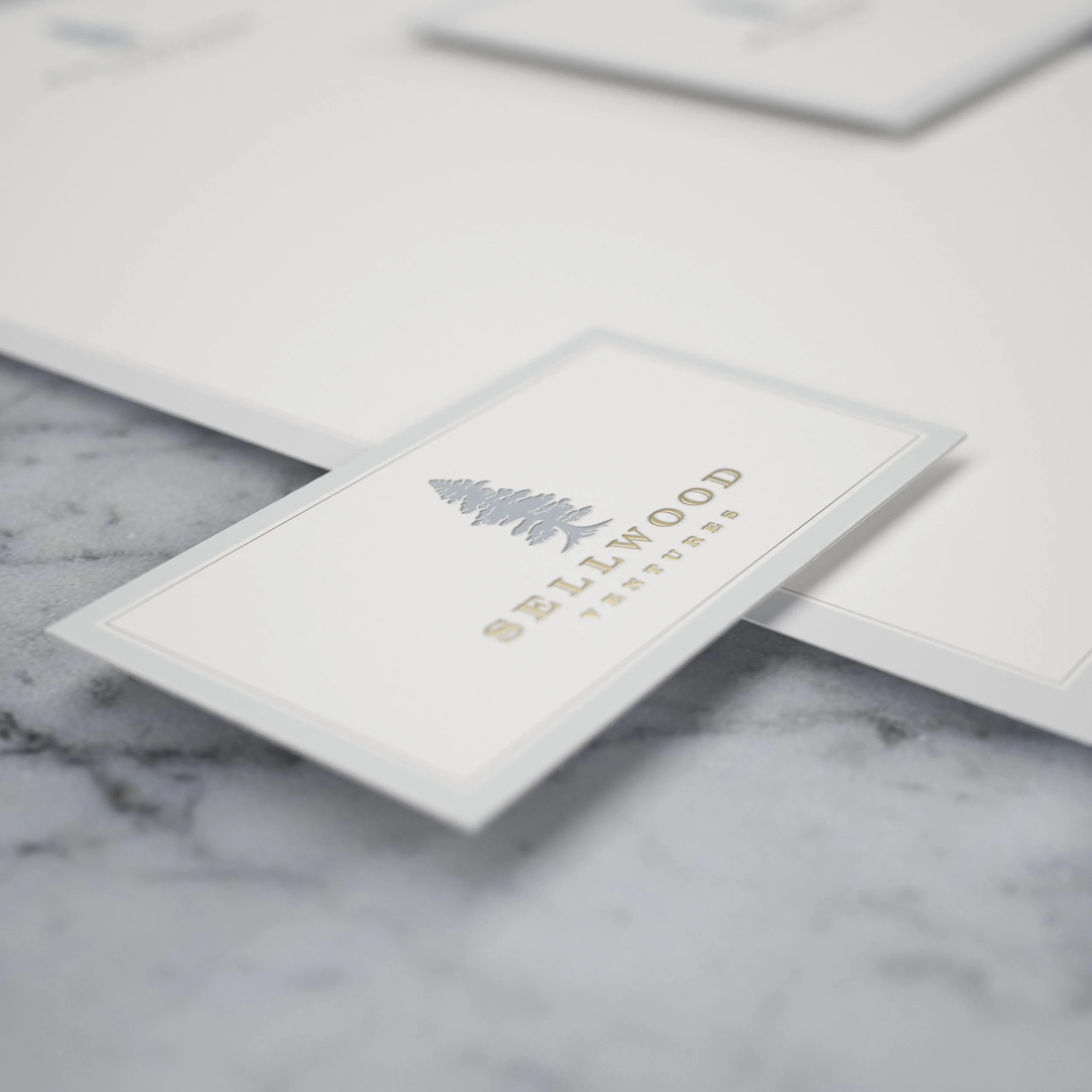 Close-up of a white card with gold lettering and a pine tree illustration, partially visible on a textured gray surface.