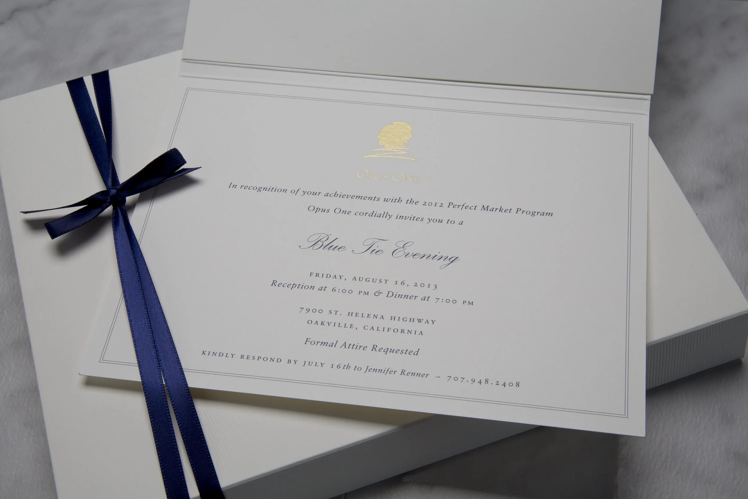 A white invitation card with a gold emblem at the top and dark blue ribbon tied in a bow on the left side. The invitation is for a formal event called the Blue Tie Evening, scheduled for August 16, 2013, in Oakville, California, with details about re
