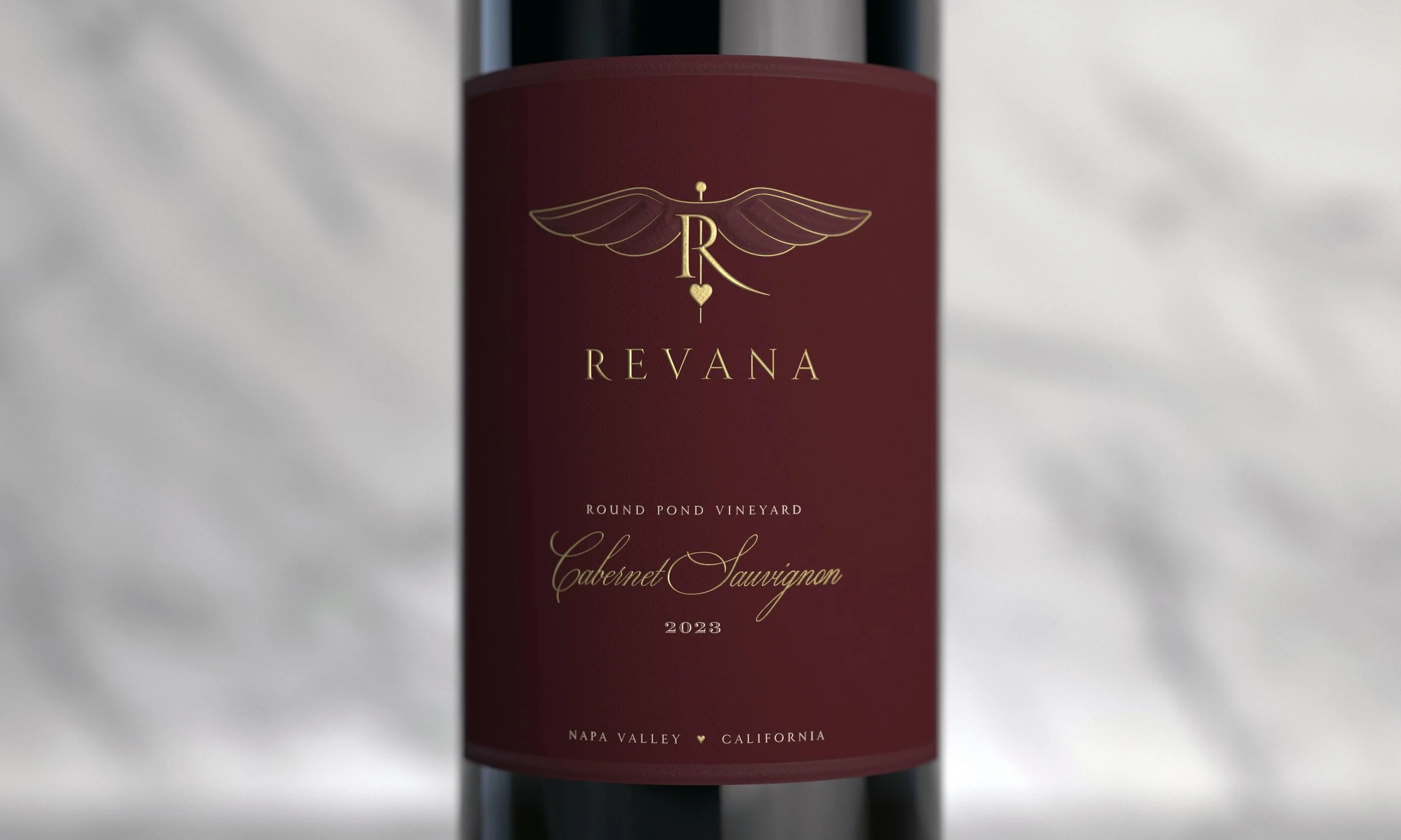 Close-up of a red wine bottle label with gold design elements, including wings and the letter R, reading 'Revana, Round Pond Vineyard, Cabernet Sauvignon 2023, Napa Valley, California.'