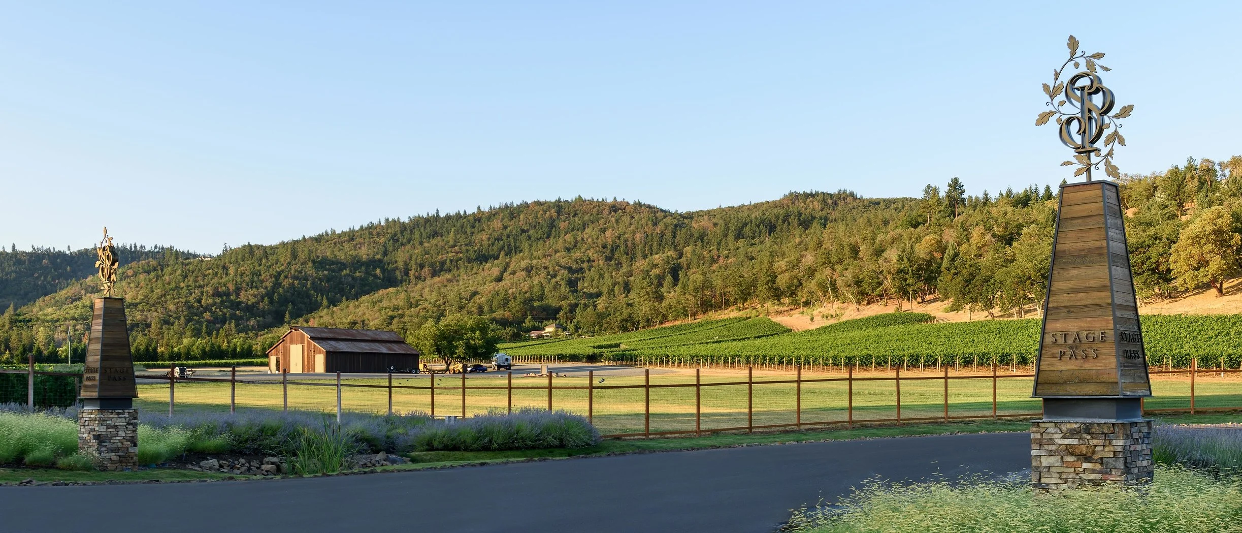 Scenic view of a vineyard with rolling hills in the background and a clear blue sky. Two tall stone pillars with metal decorative signs that say 'Stage Pass' are on either side of the image, with a wooden fence and greenery in the foreground and a sm