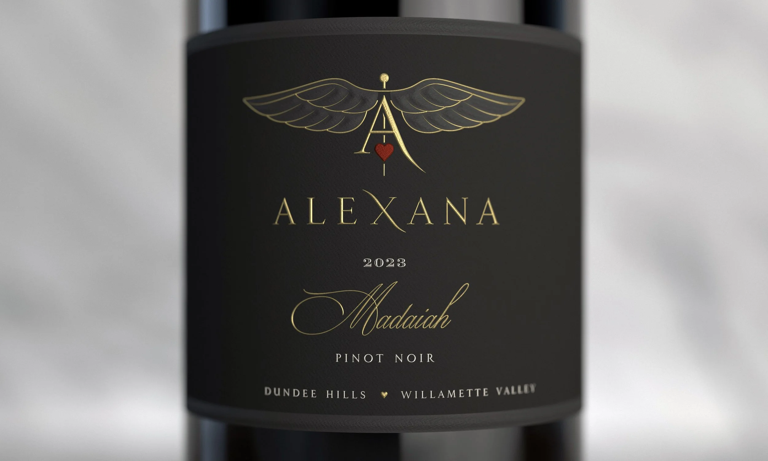 Close-up of a wine bottle label with gold and red accents. The label reads 'ALEXANA 2023 Madaiah Pinot Noir' and features a winged emblem with a heart in the center. The background is dark, and the bottle is set against a neutral backdrop.