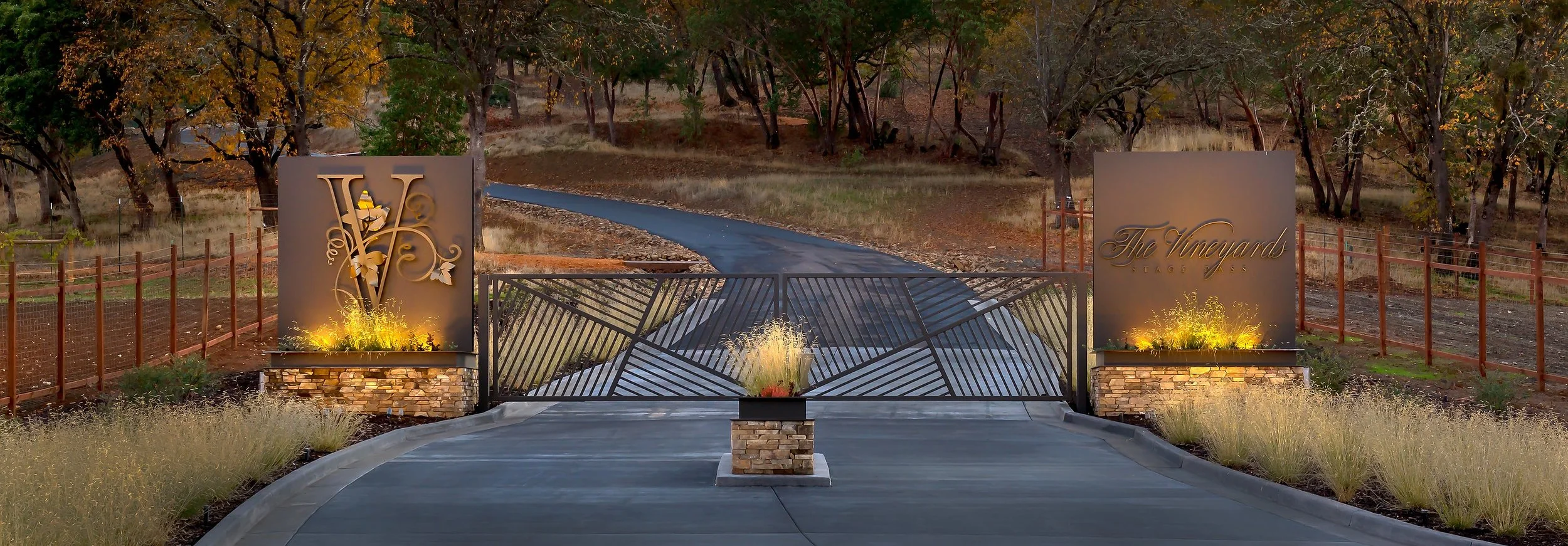 Entrance gate to The Vineyards community with decorative metal gate, two large stone signs illuminated by landscape lighting, trees with autumn foliage, and a paved pathway leading into the neighborhood.