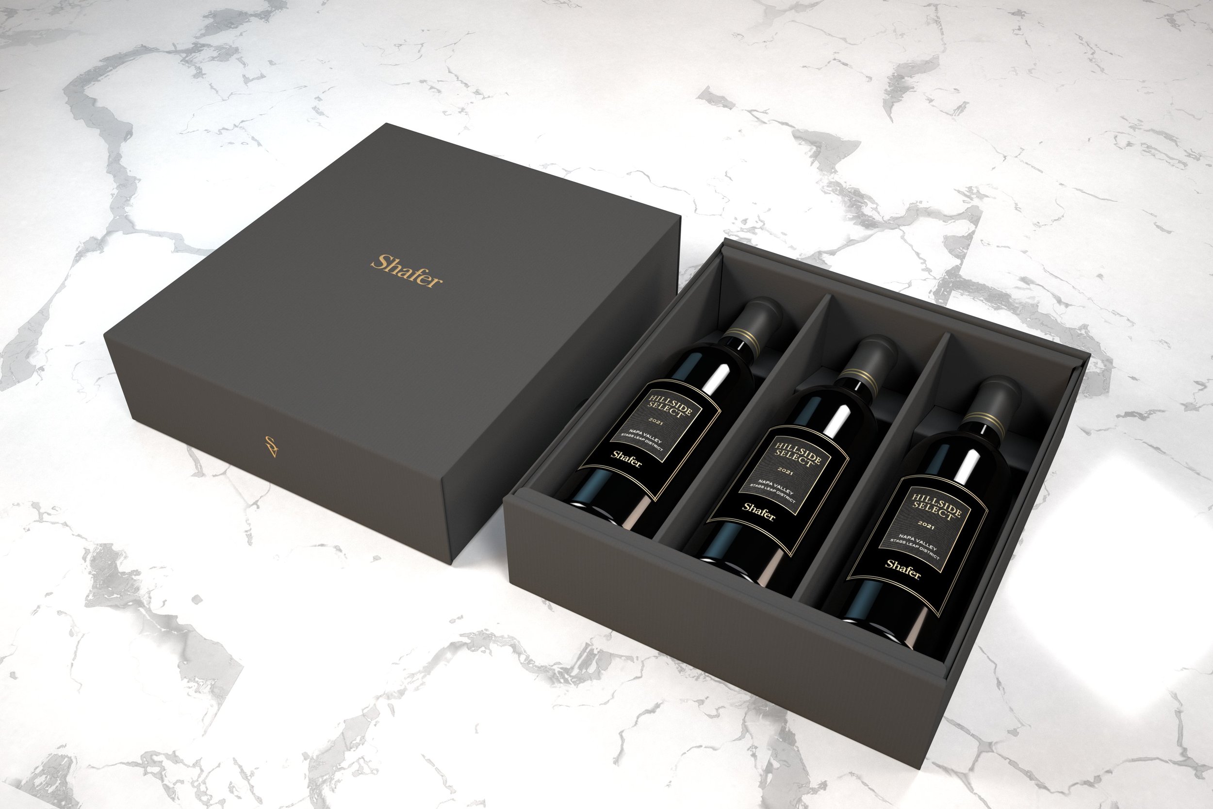 A black gift box with gold lettering that says 'Shafer' is partially open, revealing three bottles of Shafer Hillside Select wine with black labels. The box is on a white marble surface.