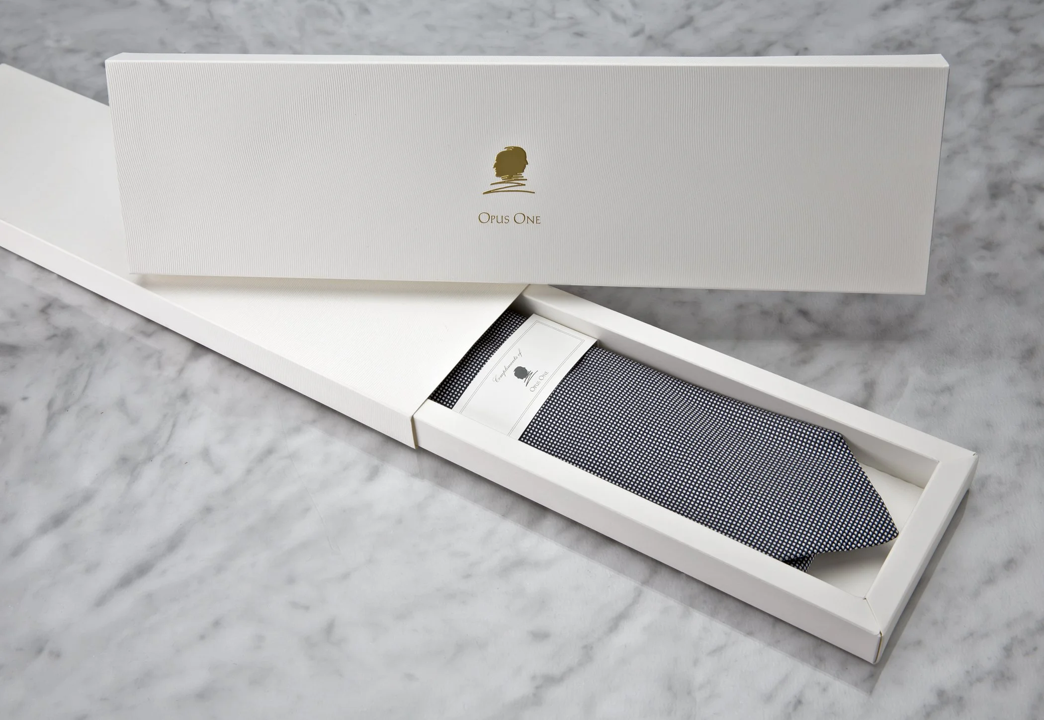 A white box with a logo of a gold profile of a person's head and the text "Opus One" underneath, placed on a white marble surface.