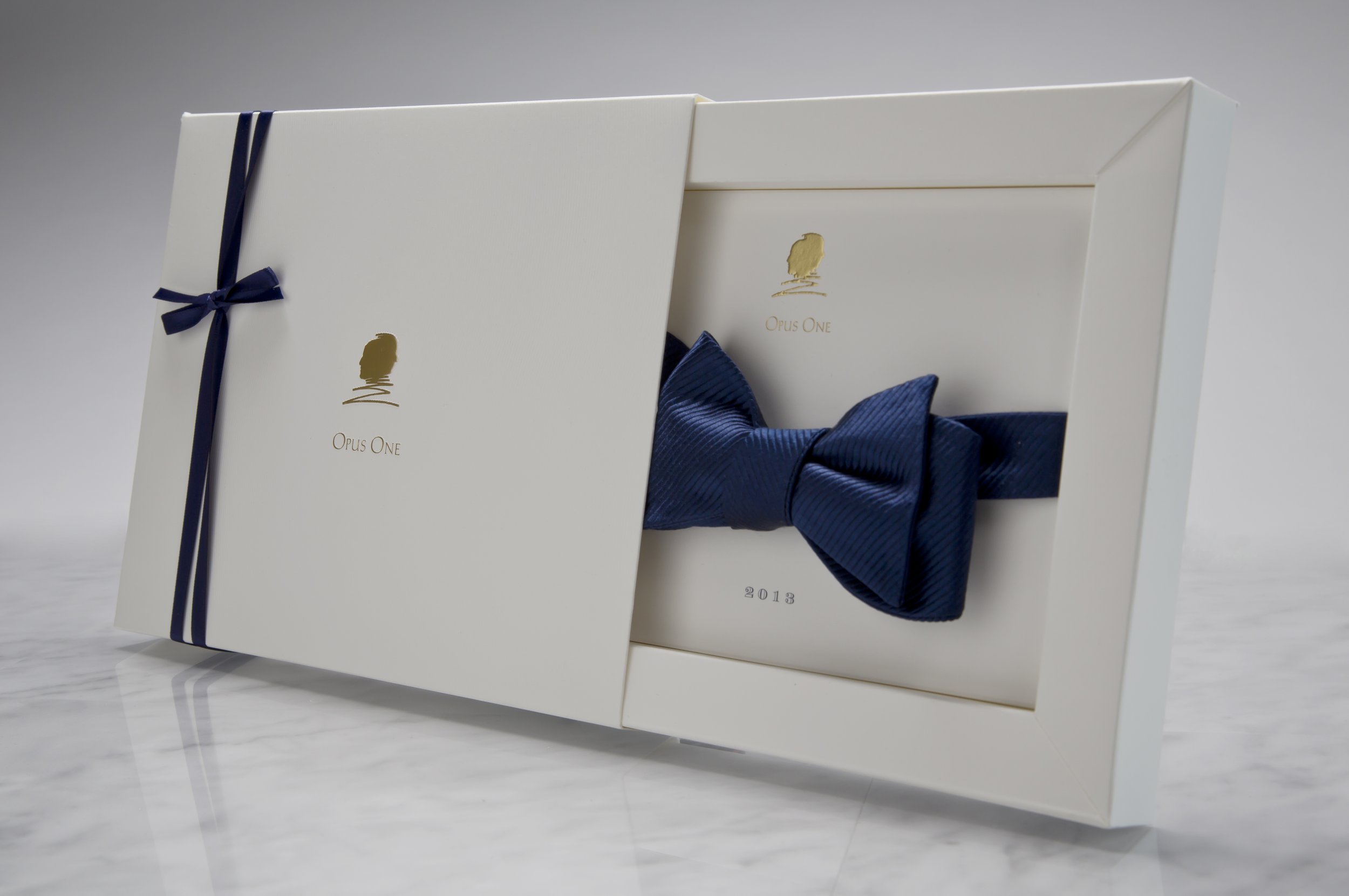 A white box with a gold logo and text, partially opened to reveal a dark blue ribbon inside, sitting on a white marble surface.