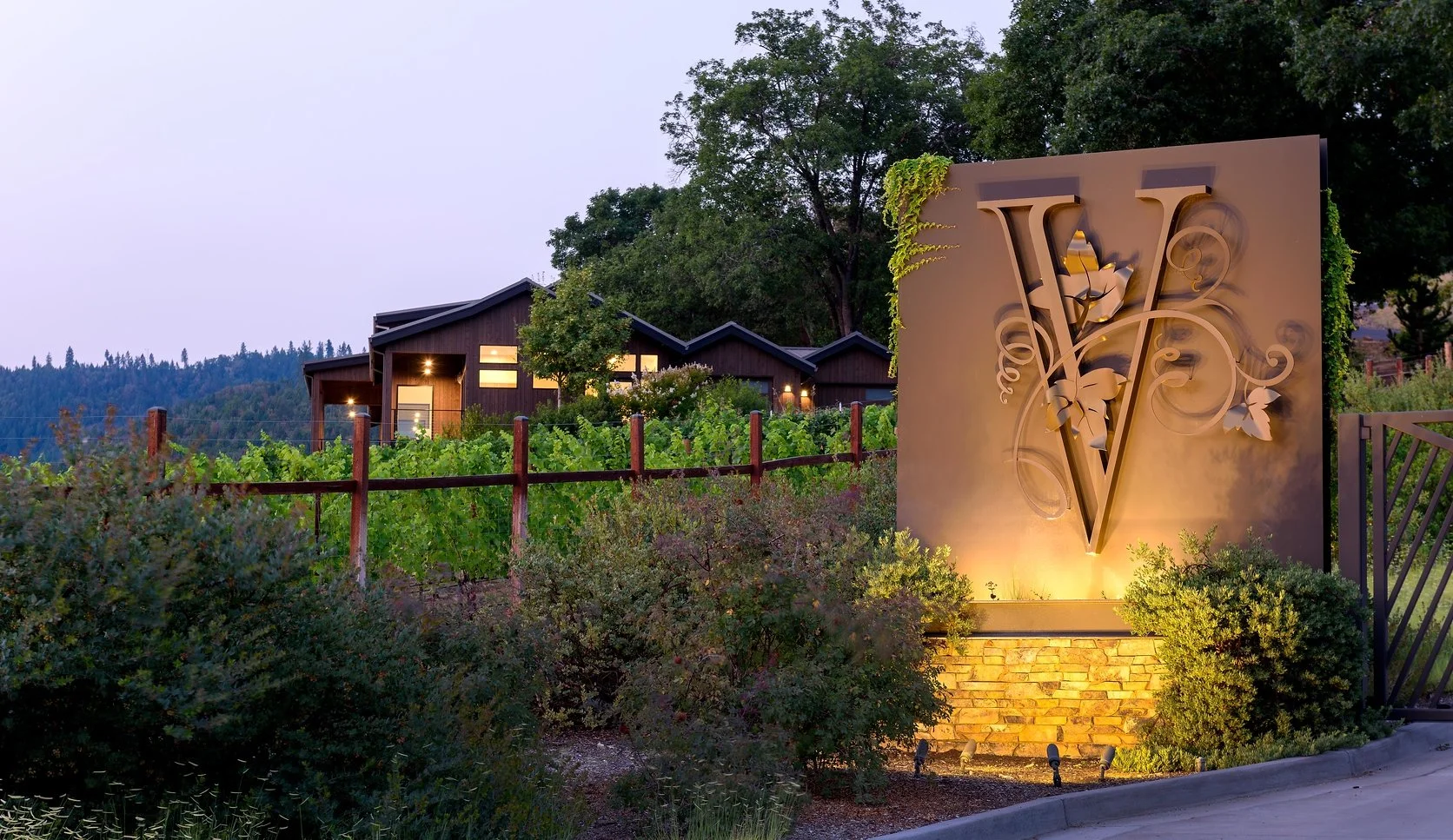 A large decorative sign with the letters V and W intertwined, featuring floral designs, illuminated at the base. In the background, there is a modern house with large windows, surrounded by green trees and a vineyard.