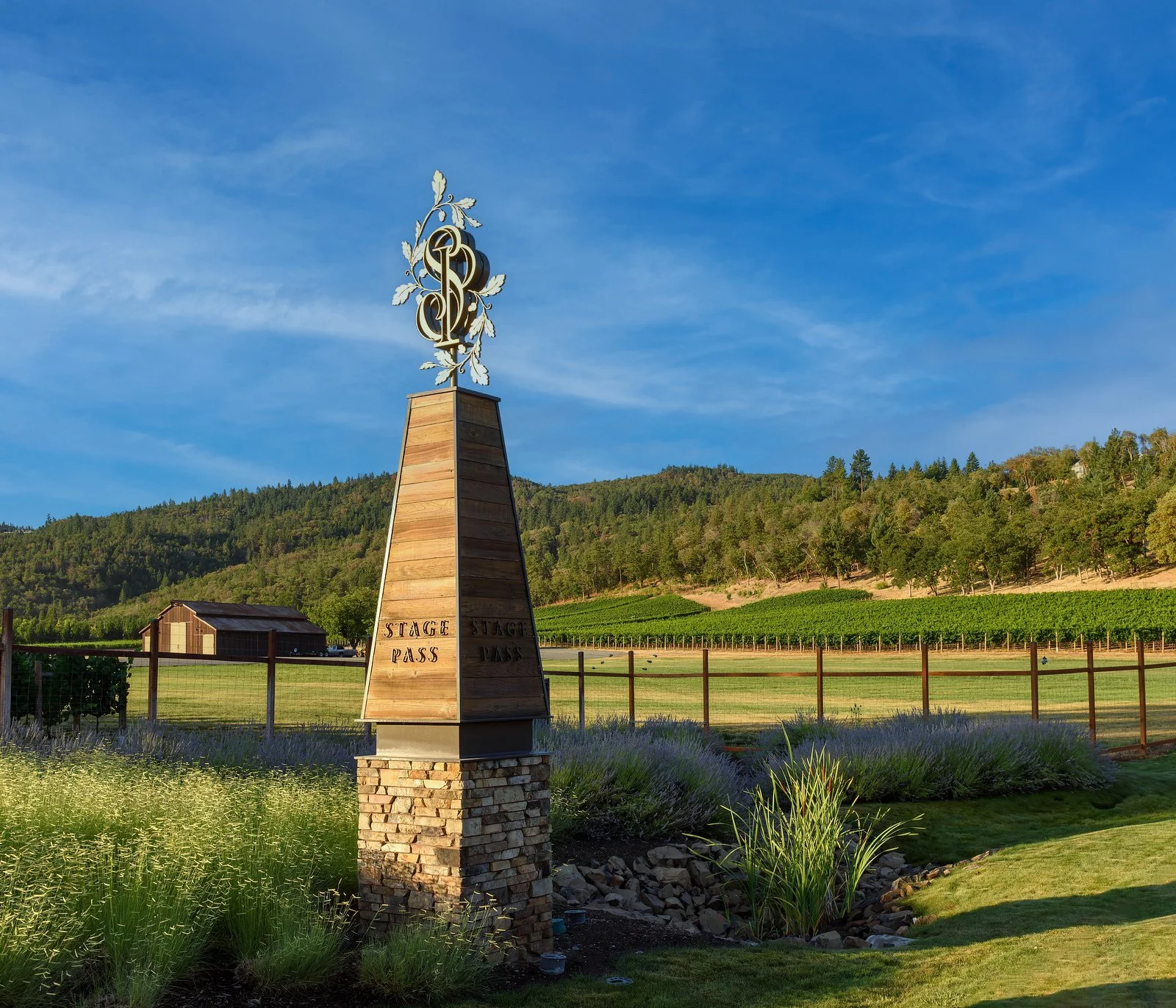 Wooden stage pass sign on a grassy area with a pond, lavender plants, a small building, rolling hills, and vineyards in the background under a blue sky.