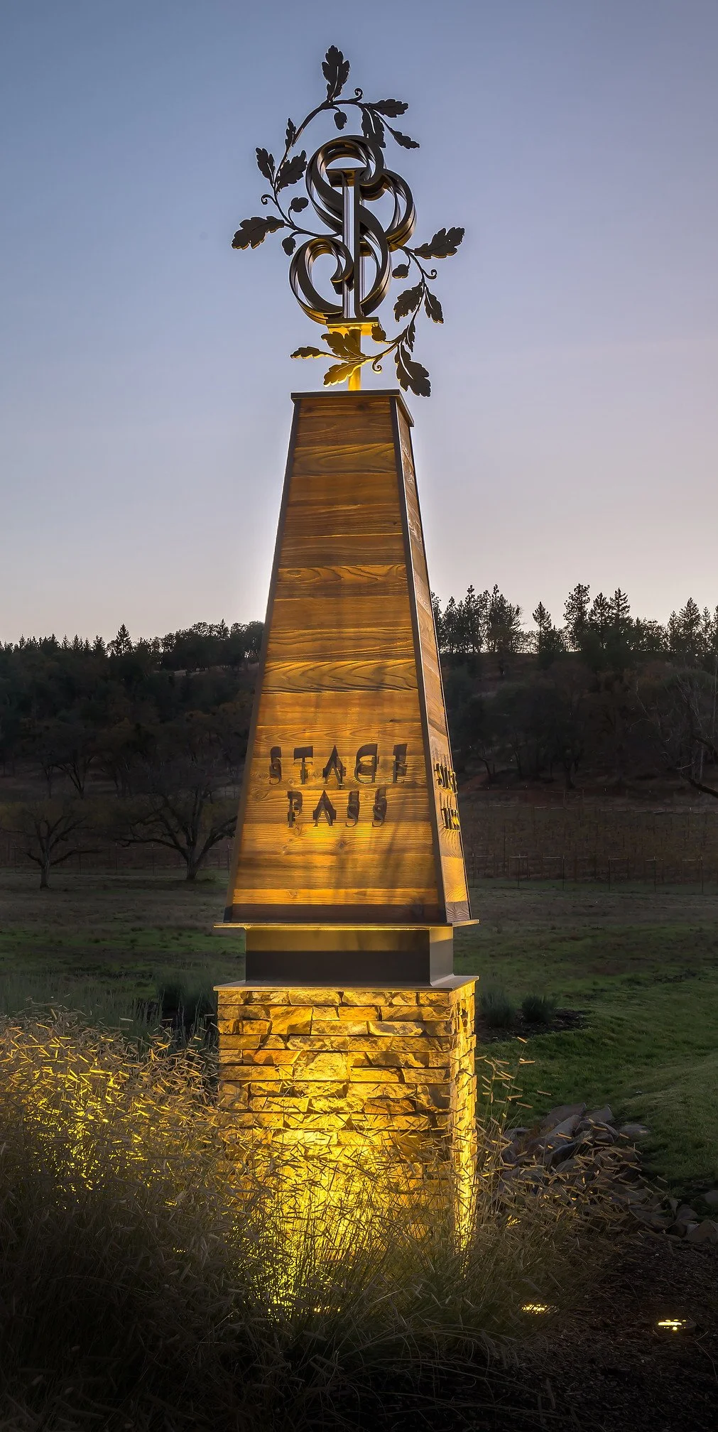 A tall decorative monument featuring a metal sculpture of a snake intertwined with a branch and leaves, situated on a wooden pedestal with inscribed text, illuminated by yellow lighting, outdoors at dusk with trees and sky in the background.