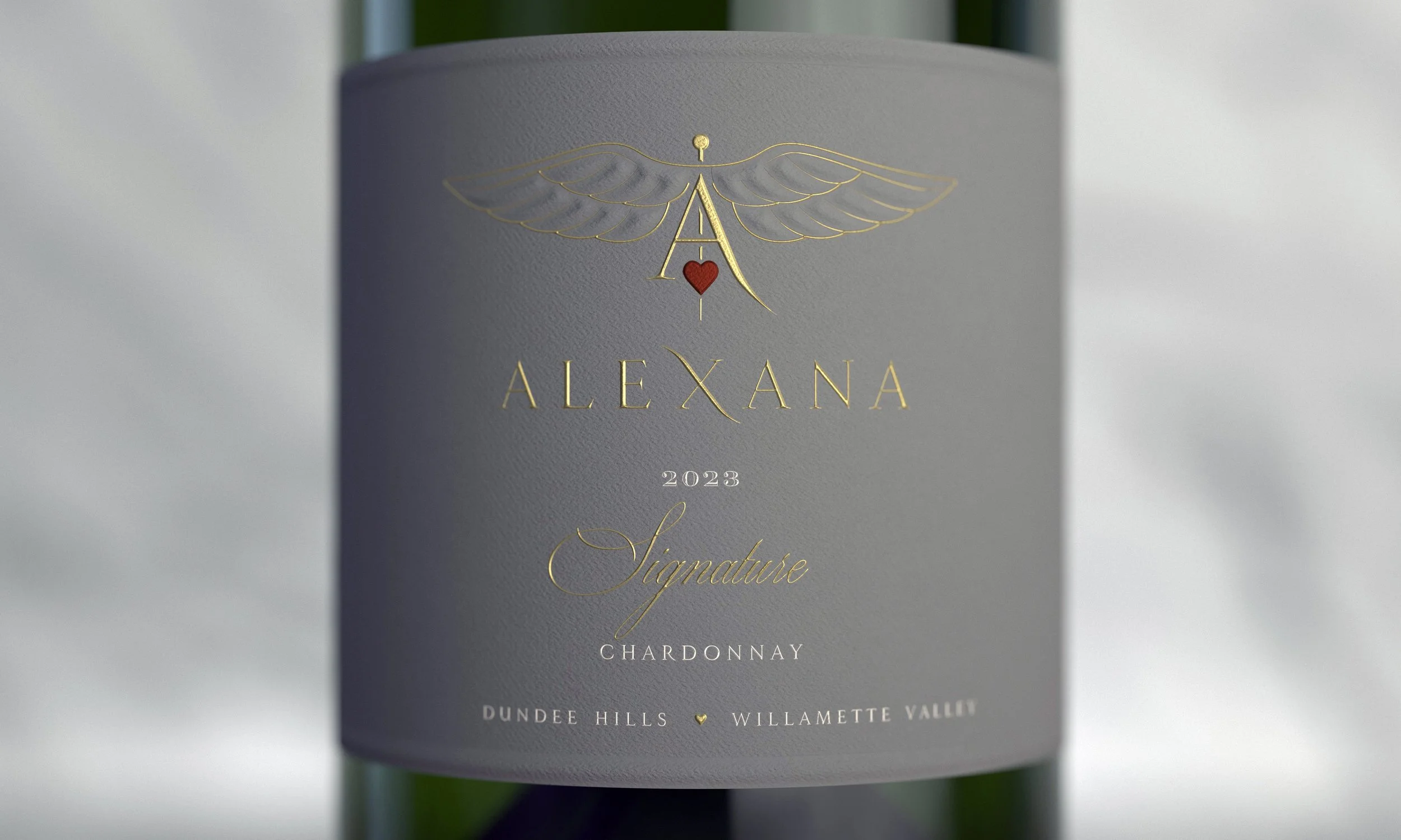 Close-up of a wine label on a bottle of Chardonnay featuring a gold winged emblem with a red heart, and the words "Alexana 2023 Signature Chardonnay" with locations Dundee Hills, Willamette Valley listed below.
