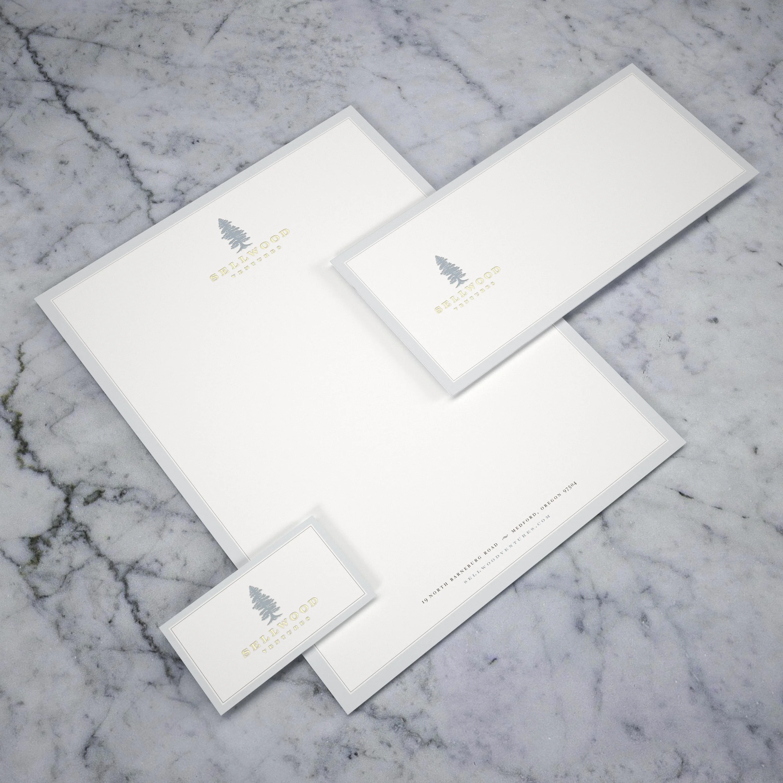 Two white rectangular cards with gold and gray text and a tree logo, placed on a white marble surface. The cards read "Selwood" and "Patios."