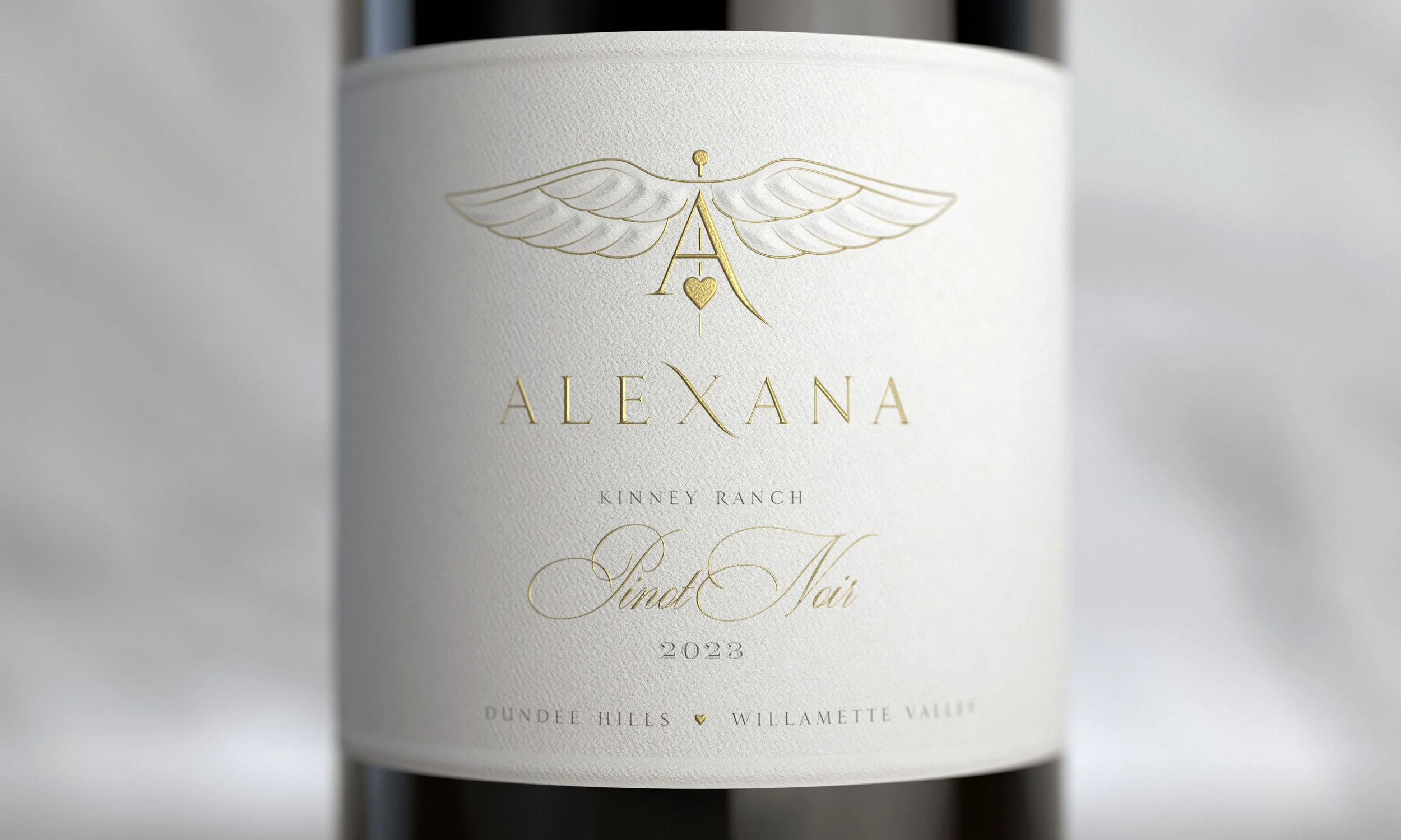 Close-up of a wine bottle label with gold and black text, including the words "Alexana" and "Pinot Noir," and a gold winged emblem with a heart at the center.