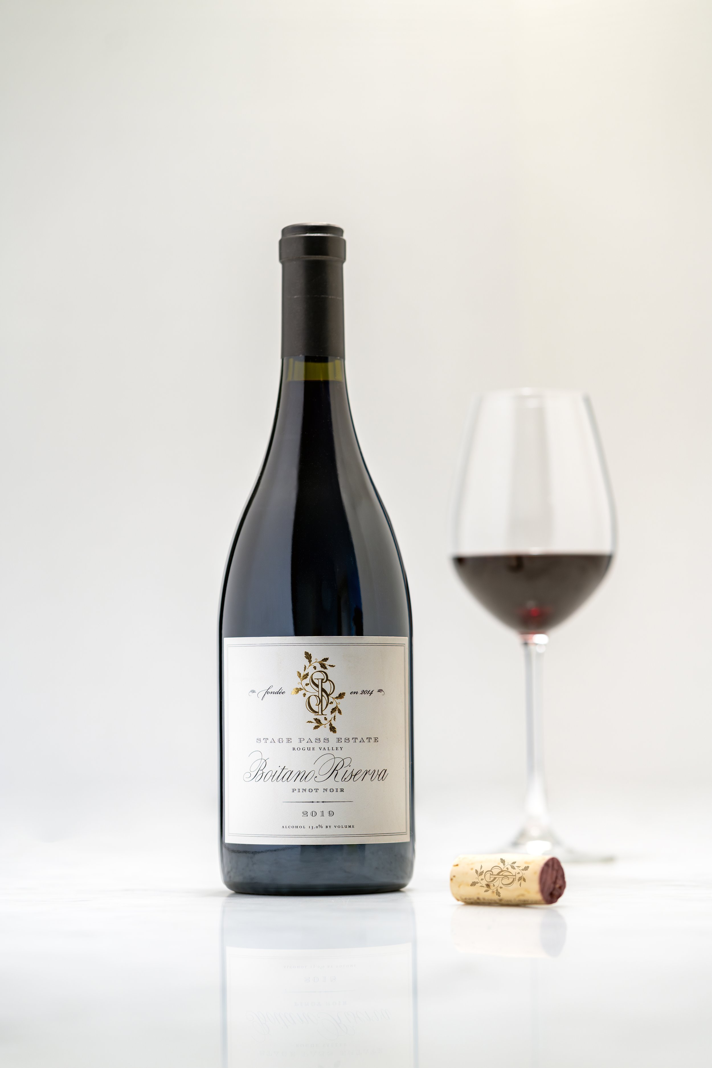 A bottle of red wine with a white label, a partially filled wine glass, and a cork on a white surface against a plain white background.