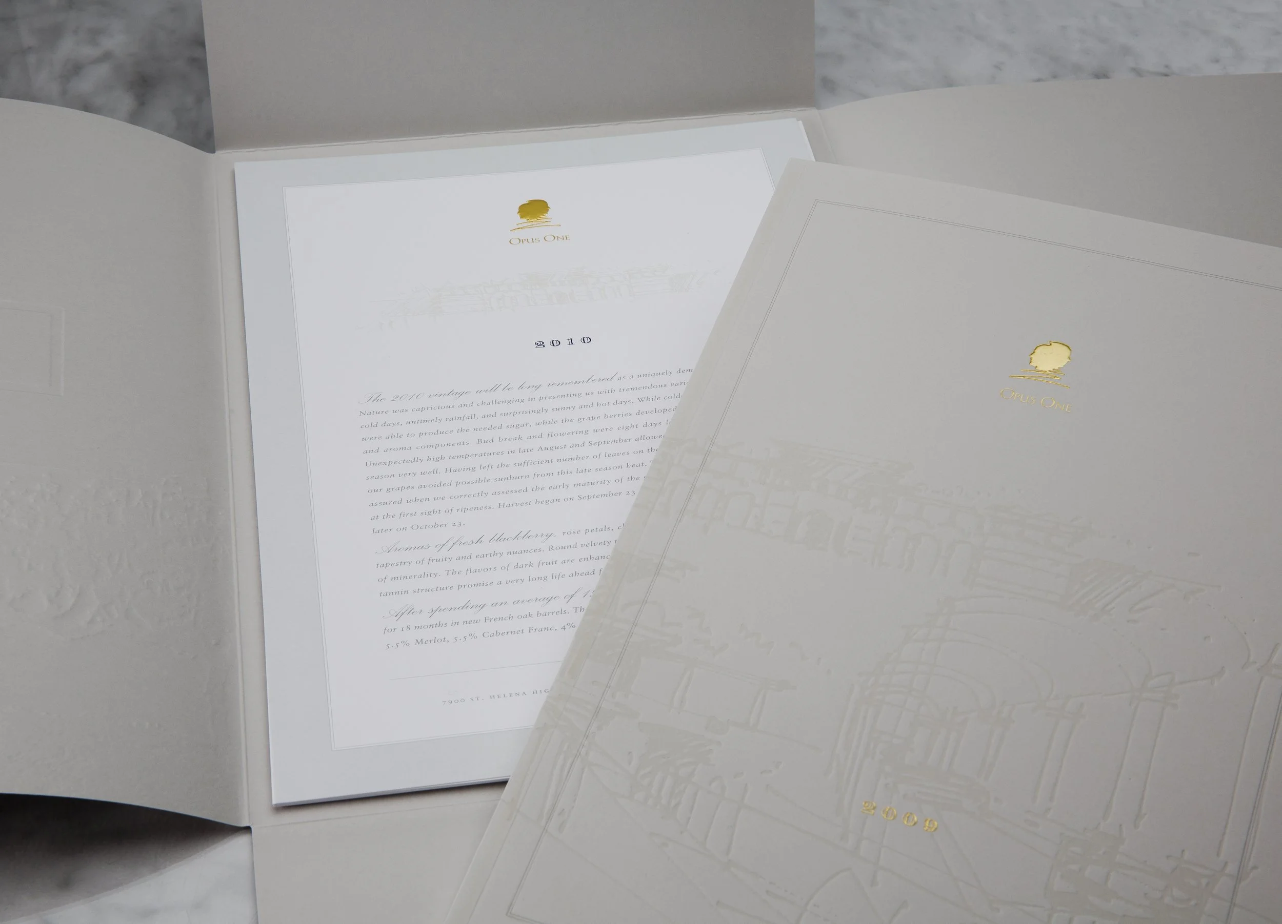A close-up of a white box with a lid, inside there is a printed brochure or program with a gold logo and the words 'Opus One' and the year '2010'. The booklet contains text, and the background is blurred with a gray, textured surface.