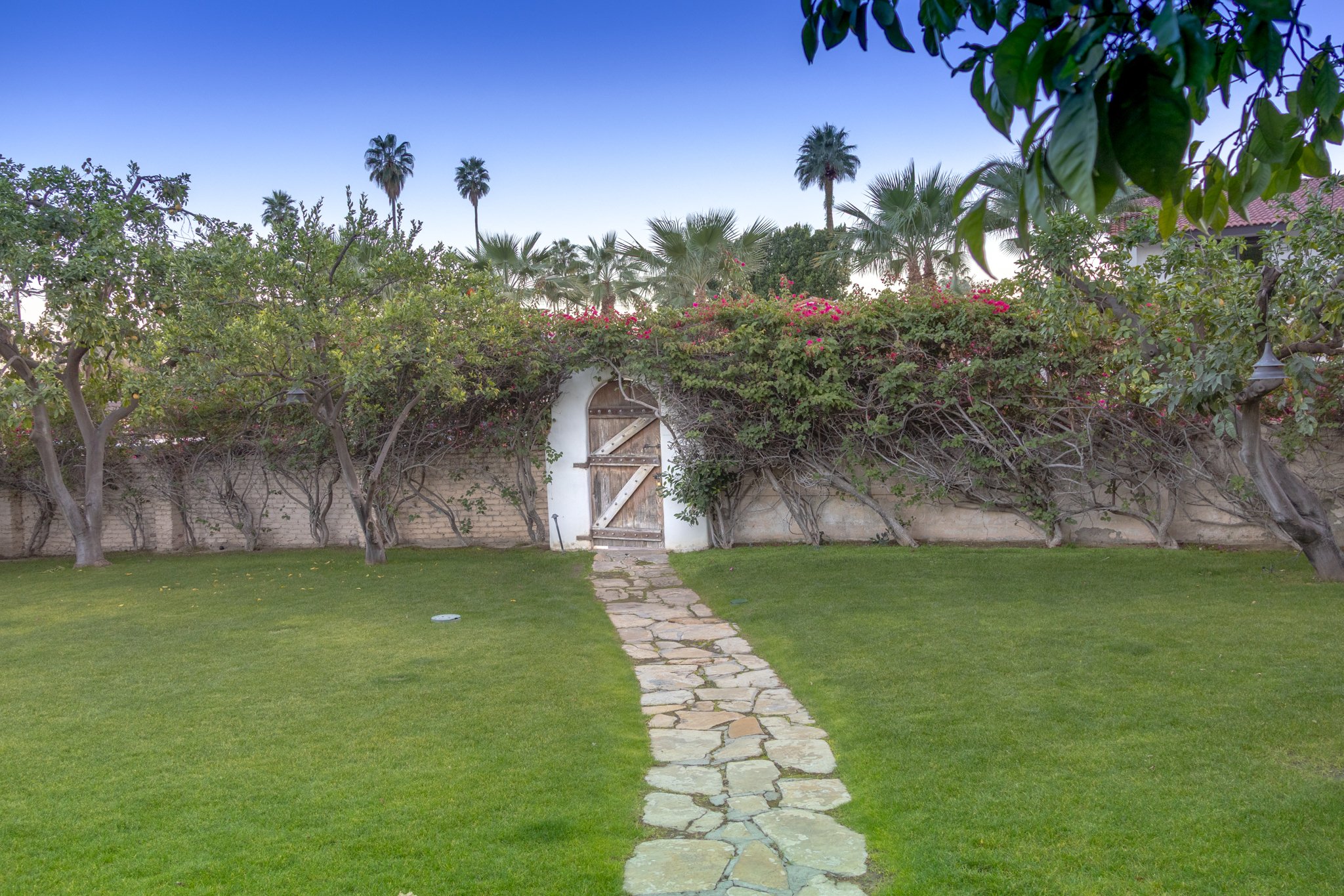 A garden with a stone pathway leading to a white wall, surrounded by trees and bushes, with a wooden gate and pink flowers, under a clear blue sky in the background.