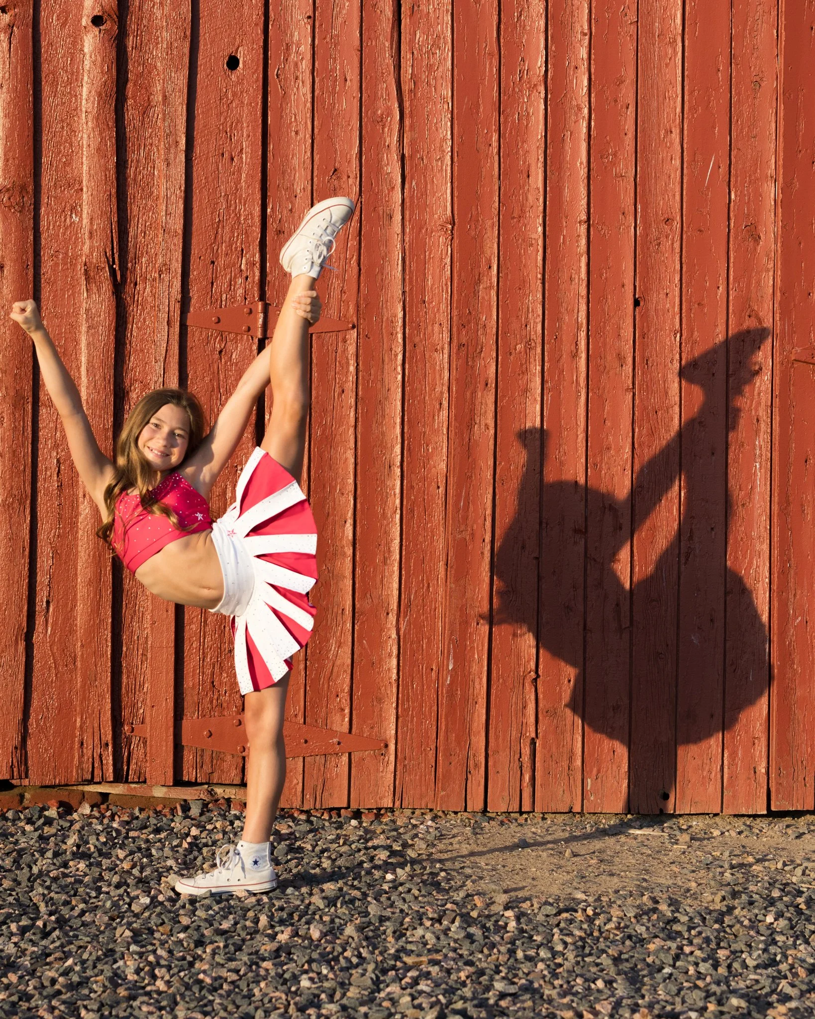 A portrait of a young girl in a cheerleader outfit standing on one leg, raising her arms, and smiling. She is positioned in front of a large red wooden gate, with sunlight casting a shadow of her on the gate, and small rocks on the ground.