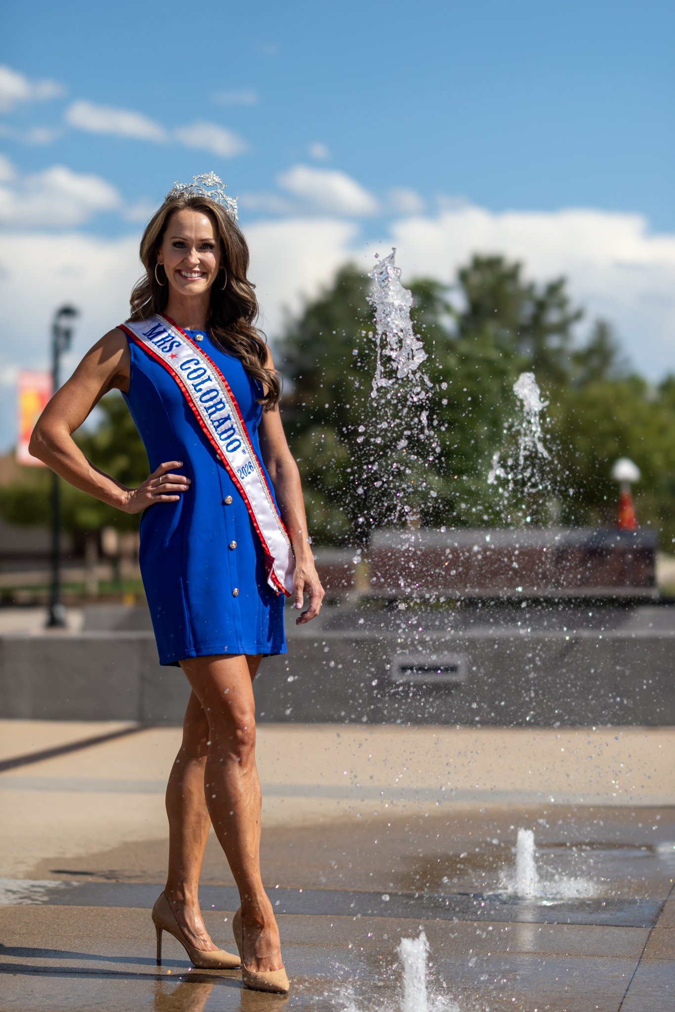 A portrait of a woman standing on a street near a fountain, wearing a blue dress, high heels, a sash that reads 'Miss Colorado 2023,' and a tiara, smiling at the camera with water splashing nearby.
