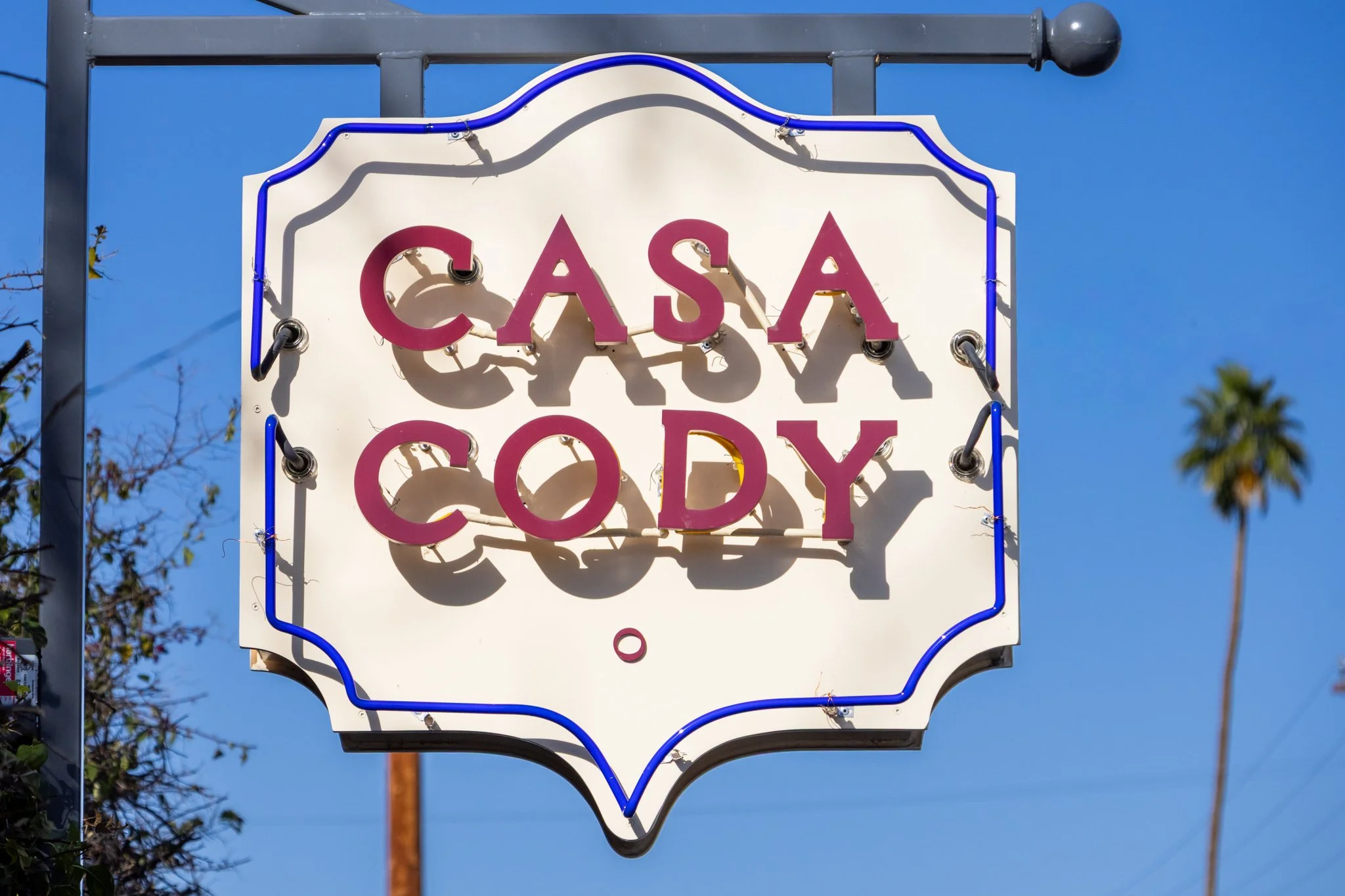 A neon sign reading 'Casa Cady' in pink letters, mounted on a white background with a blue outline, against a clear blue sky.