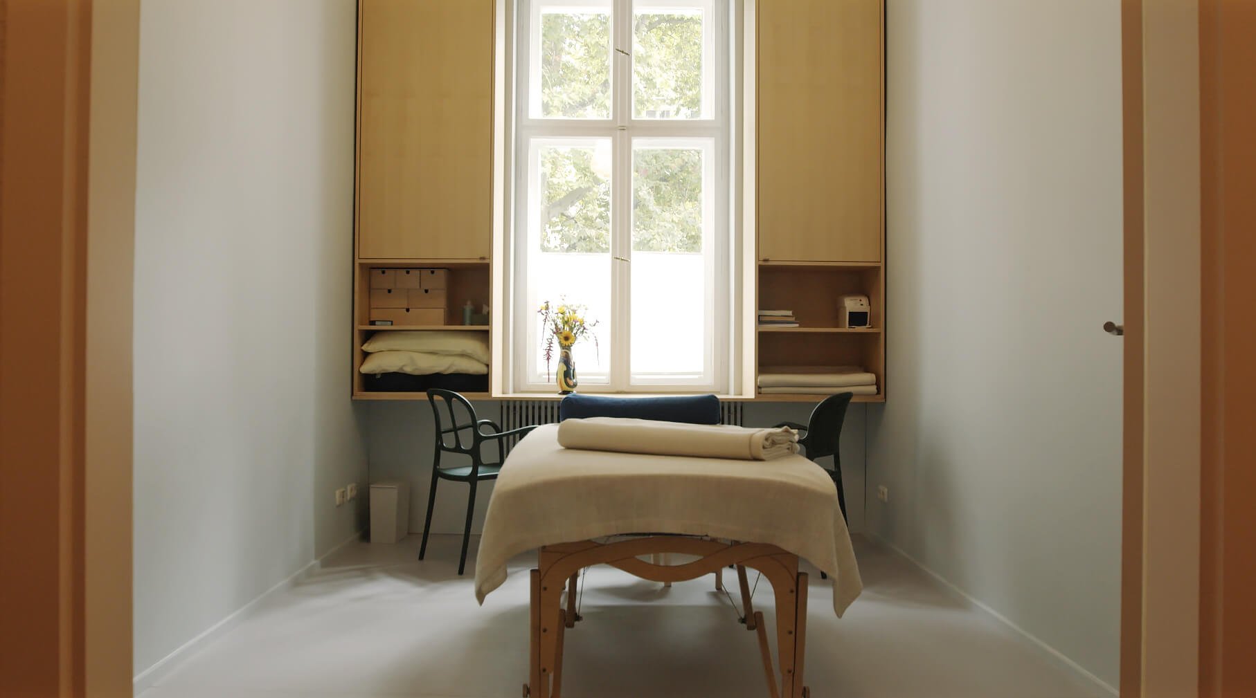 Behandlungsraum mit Tisch, Stühlen, Fenster mit Tageslicht, und Regalen an der Wand.