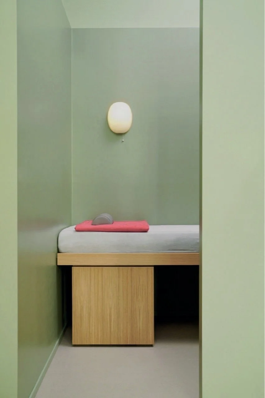Ein Behandlungsraum mit einem Bett, das eine rosa Decke und eine graue Kopfstütze hat, an der Wand hängt eine weiße Scheinlampe.
