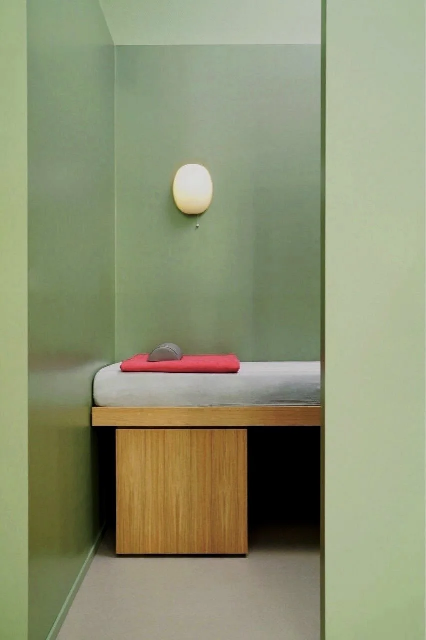 Ein kleines, minimalistisches Behandlungszimmer mit einem Einzelbett, einem roten Handtuch, einer weißen Lampe an der Wand und grünen Wänden.