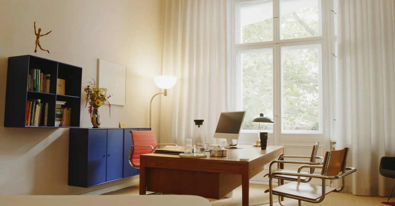 Ein komfortabler Arbeitsbereich mit einem großen Holzschreibtisch, einem Computer, einer Schreibtischlampe und mehreren Stühlen vor einem großen Fenster mit weißen Vorhängen. Im Hintergrund befindet sich eine Wandregal mit Büchern, einer Vase mit Blumen und einer Stehleuchte.