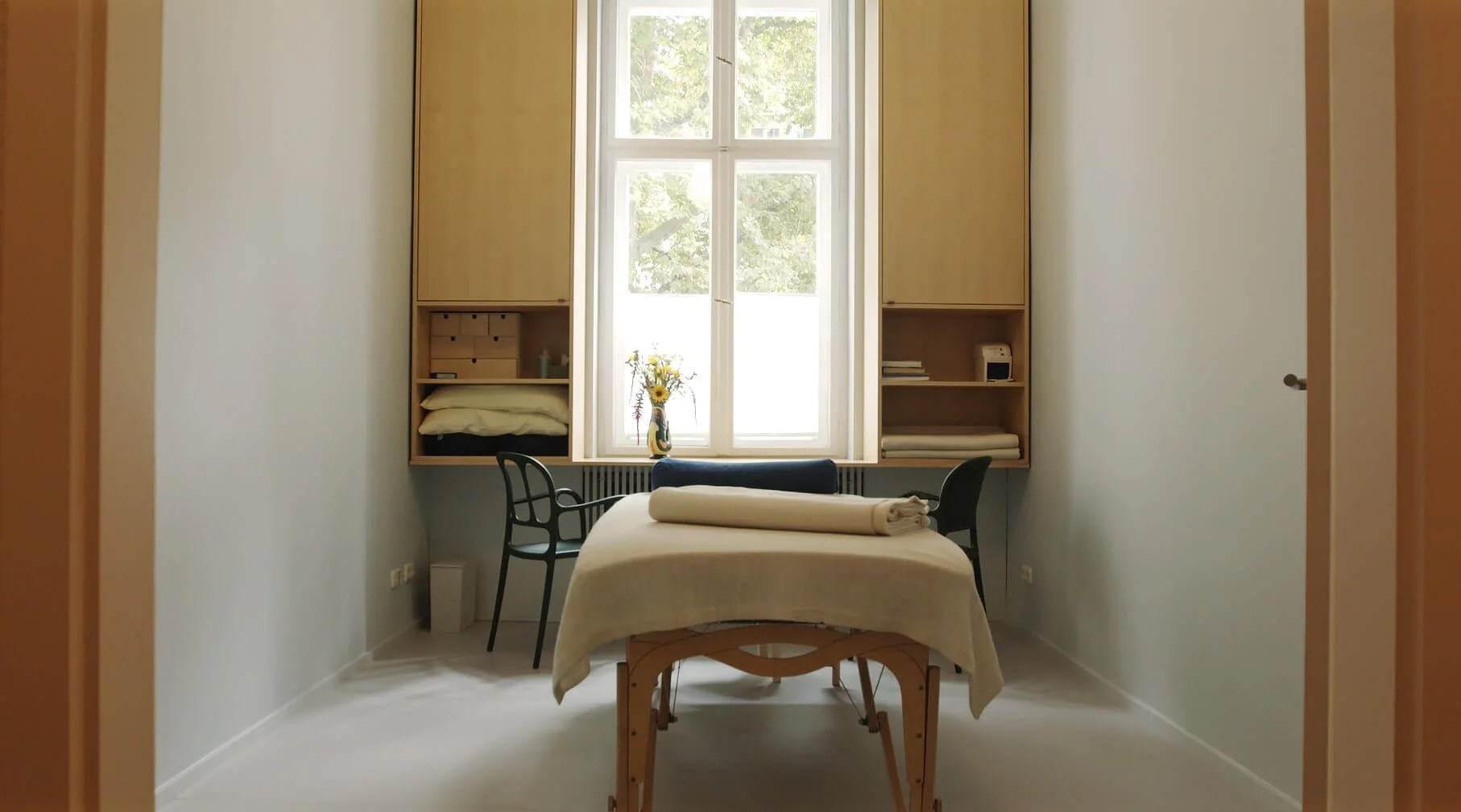 Behandlungsraum mit Therapietisch, zwei Stühlen, großen Fenster mit Blick nach draußen, Bücherregal mit Büchern, Handtüchern und Dekorationen, Sonnenblumen in Vase.
